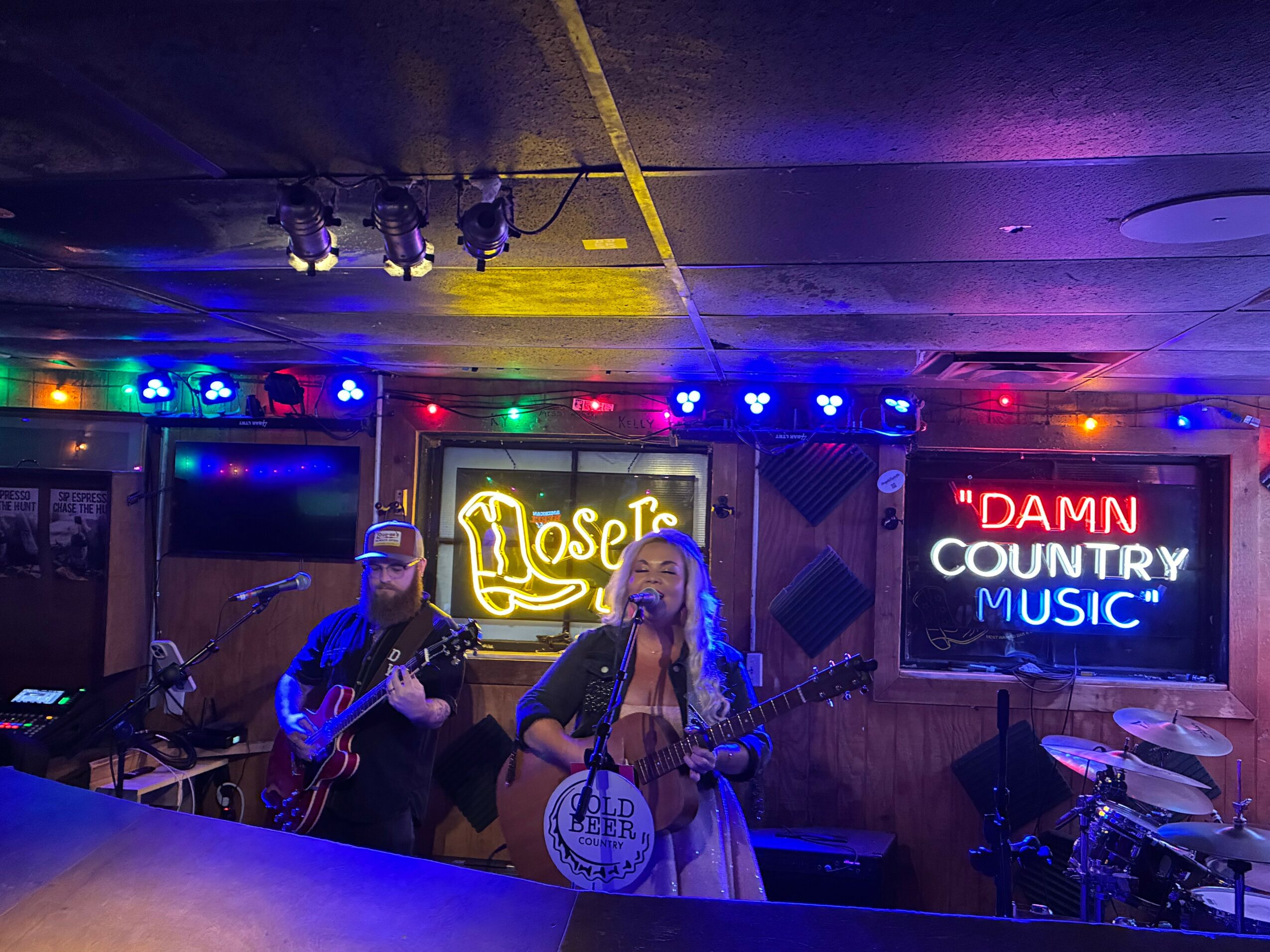 Two musicians perform on stage with colorful lights, a neon sign, and a drum set in the background.