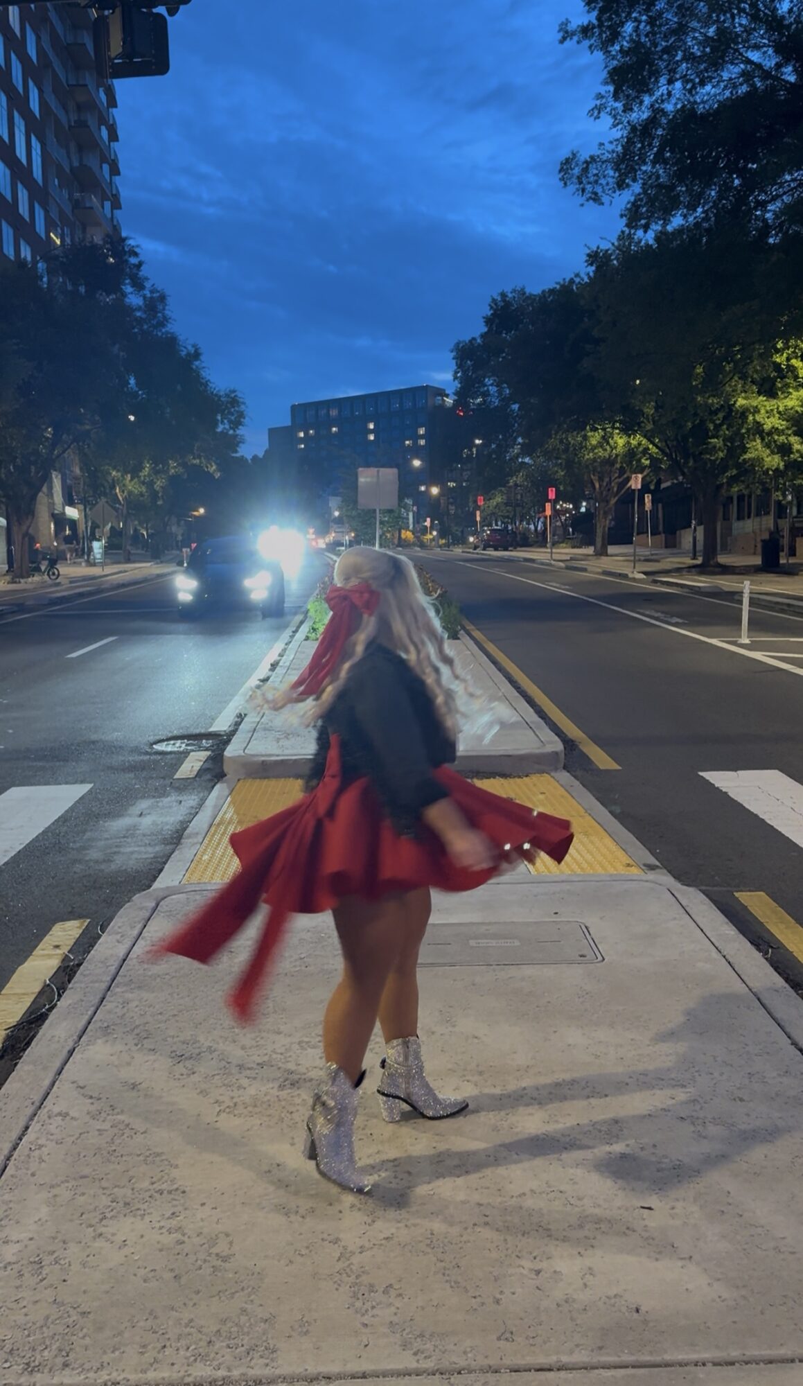 Person with long hair and a red dress walking on a city street at dusk, trees and buildings in background.