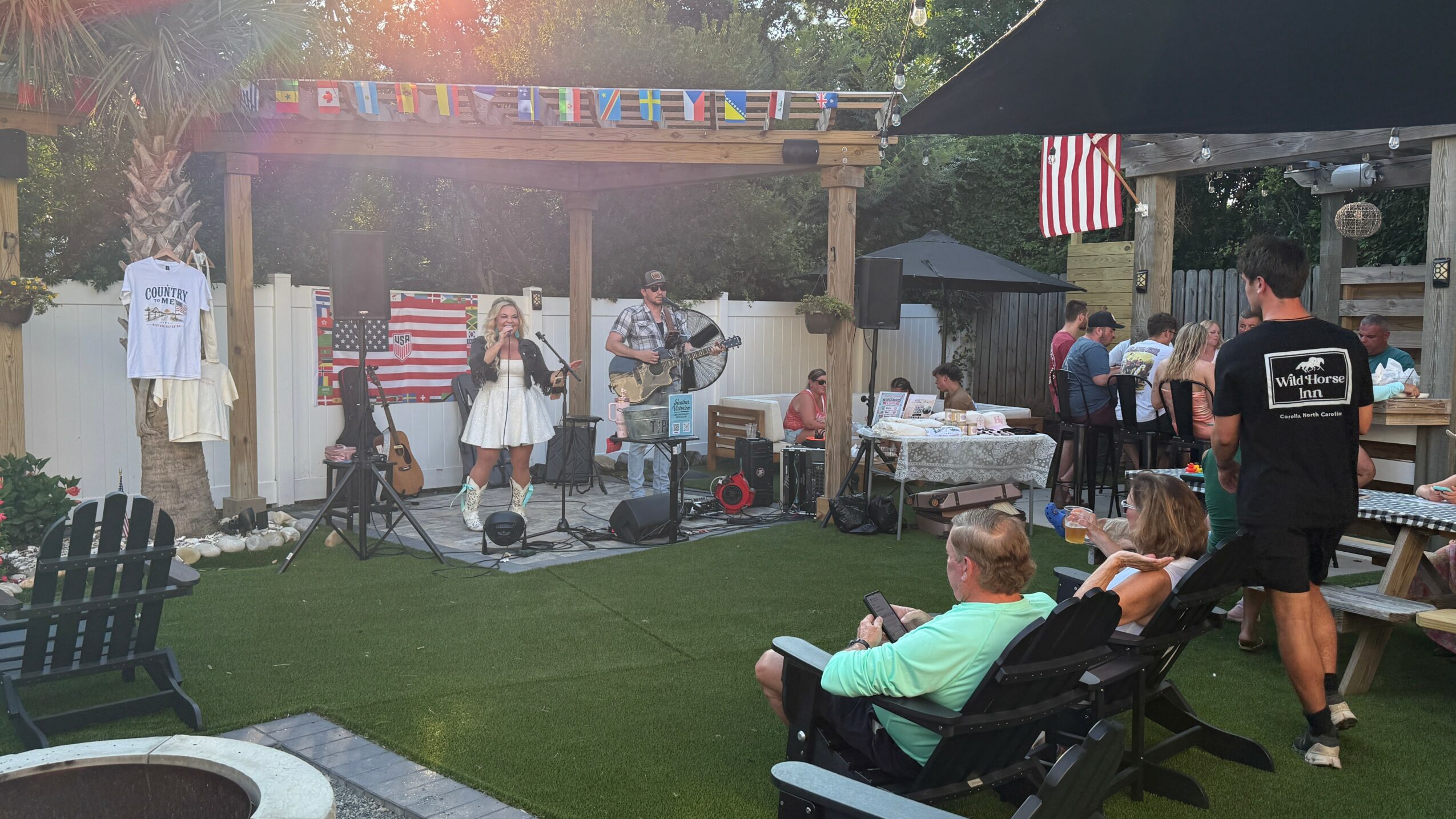 People perform music on a small outdoor stage decorated with American flags, with audience seated on benches and chairs.