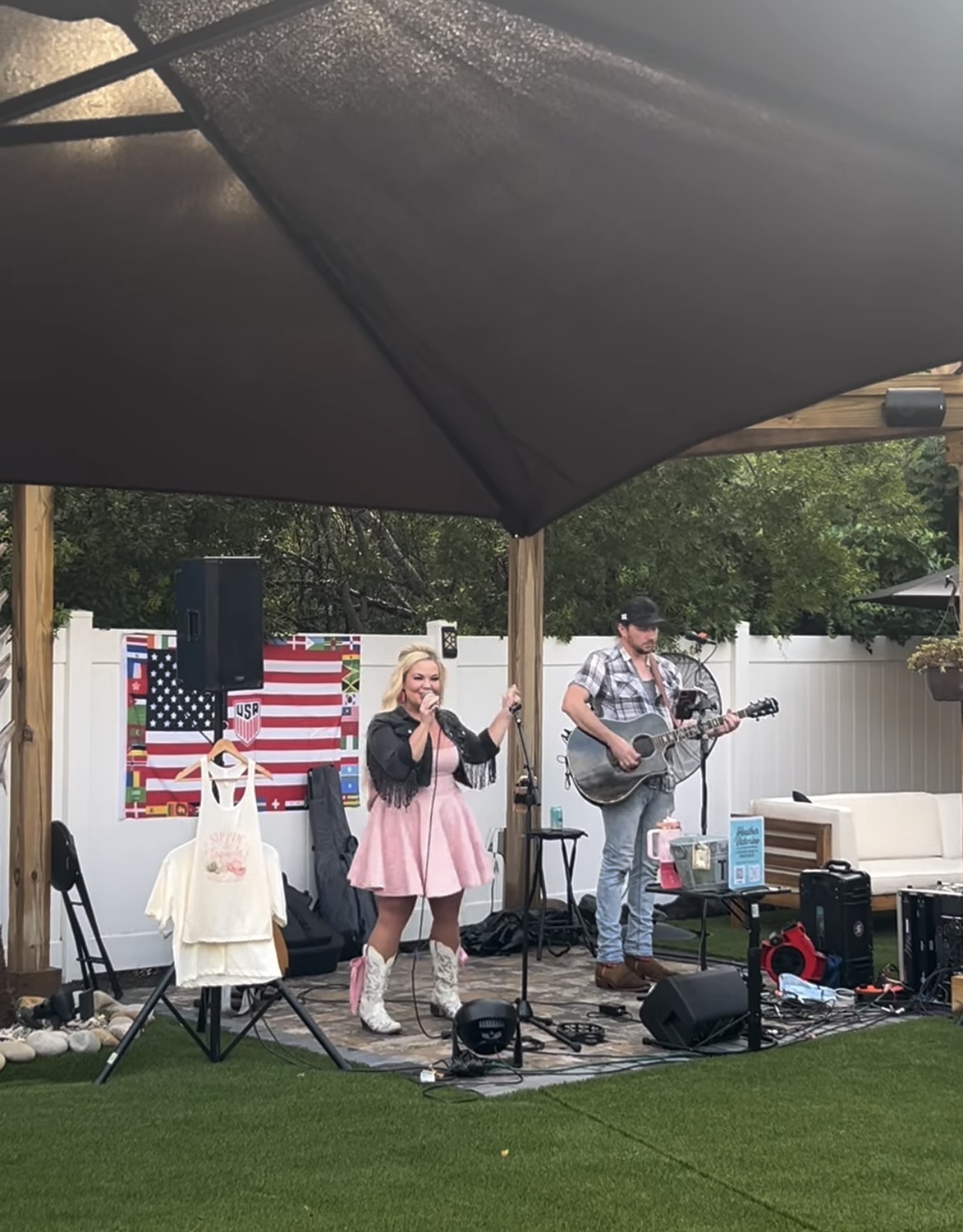A girl singing and a boy playing guitar on an outdoor stage with an American flag backdrop, under a large canopy.