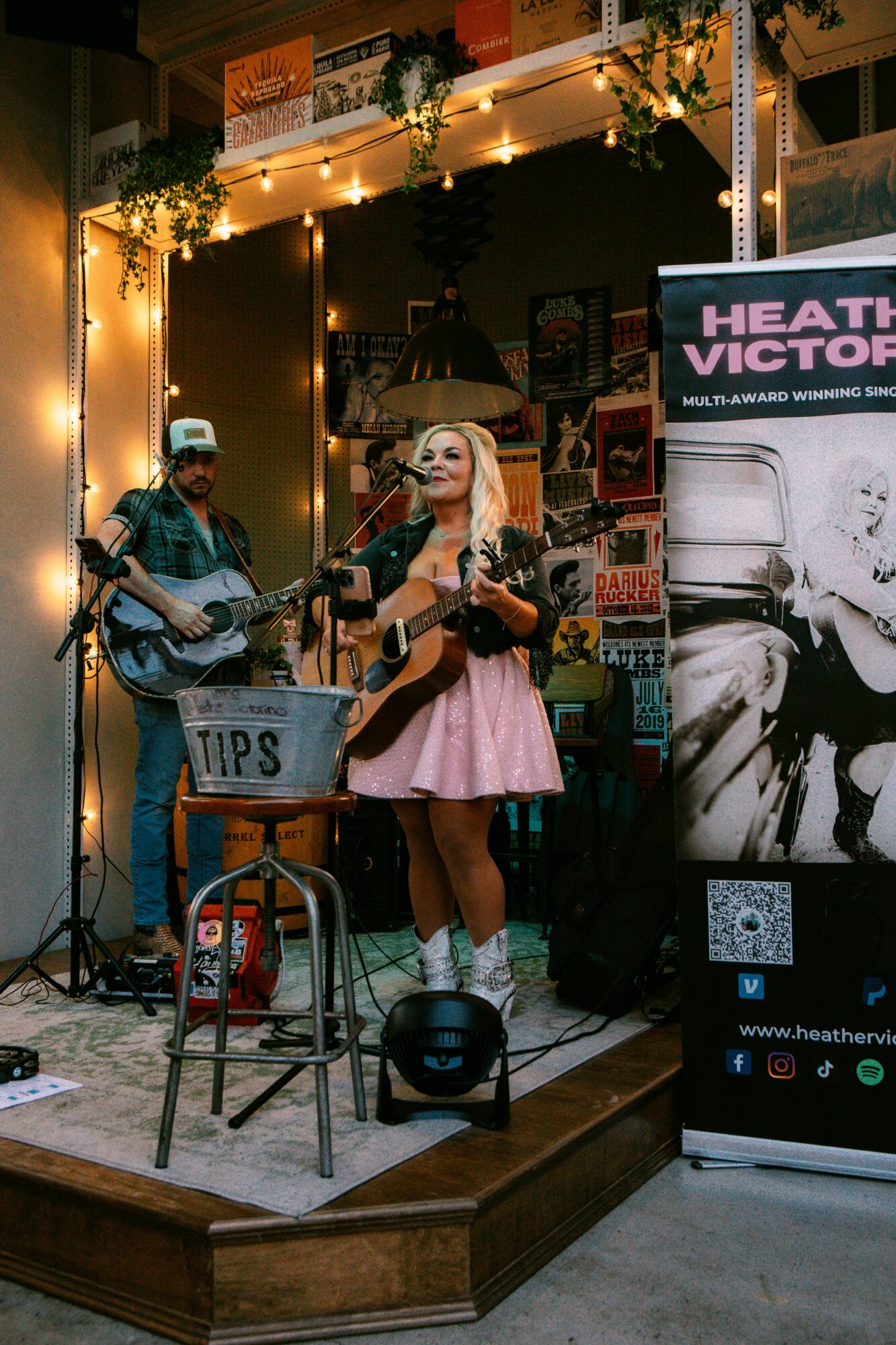 Two musicians perform on a small stage with a woman singing and a man playing guitar, illuminated by warm lighting.