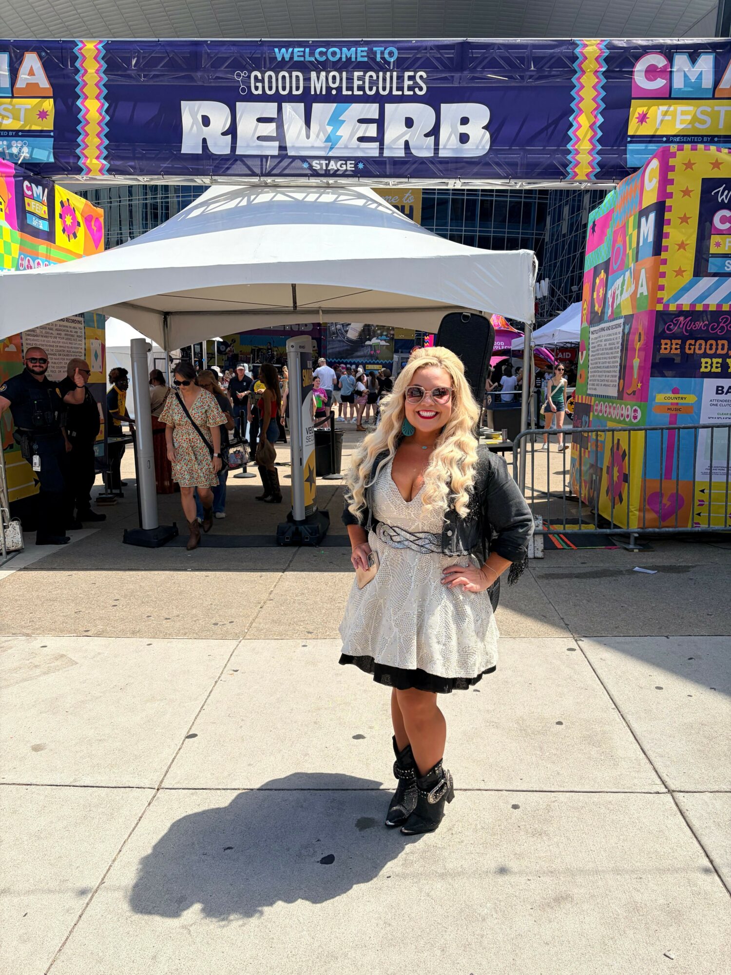 A person with long blonde hair, wearing a white dress and black boots, stands outside a booth at a fair or event.