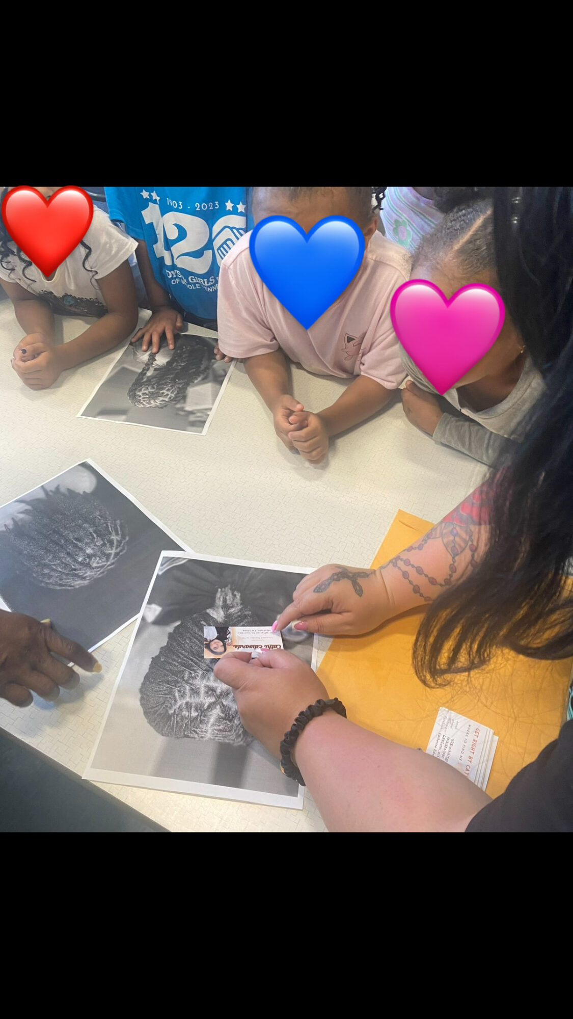People gathered around a table looking at photographs, with faces covered by colored hearts, in a well-lit room.