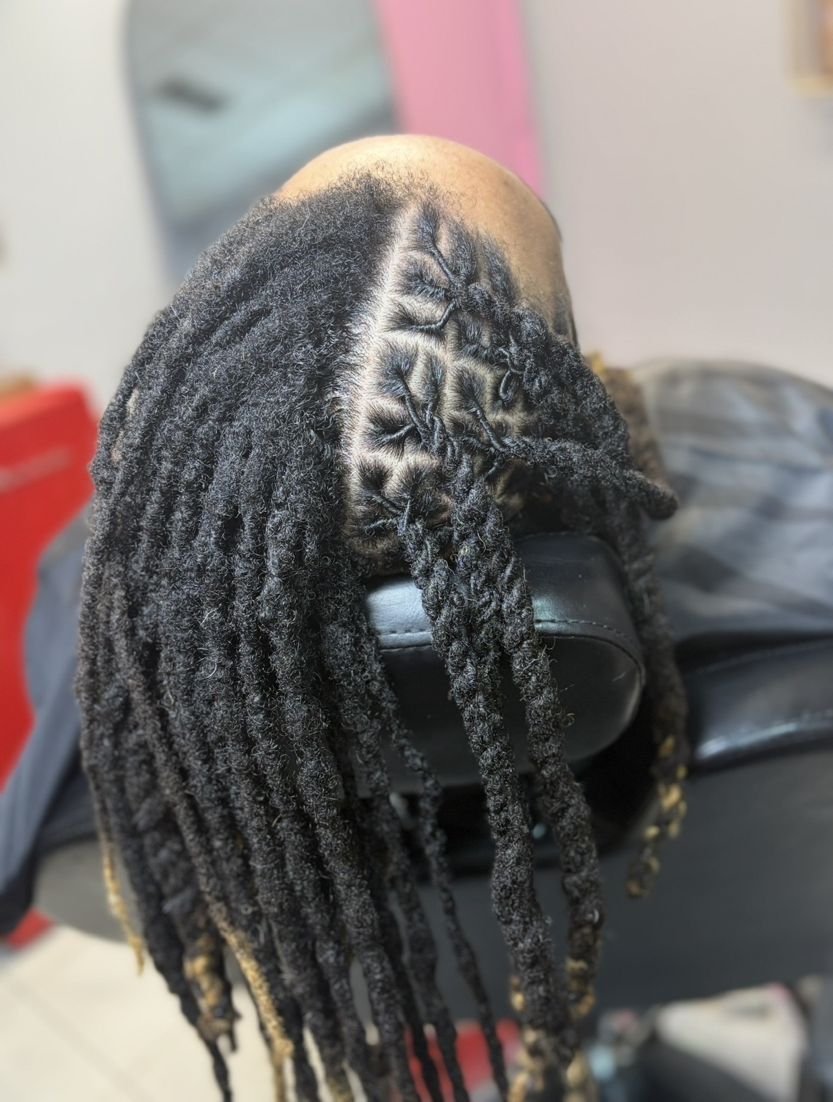 Person with dreadlocks resting head on a black surface, background includes a mirror and a pink wall.