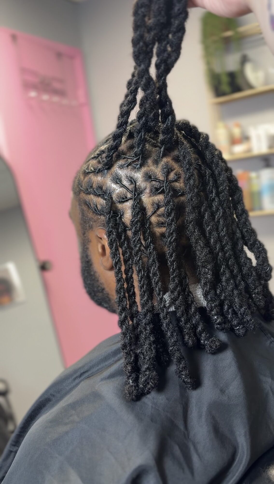Person with braided hairstyle, holding a section of hair up, in a room with pink door and shelves, viewed from behind.