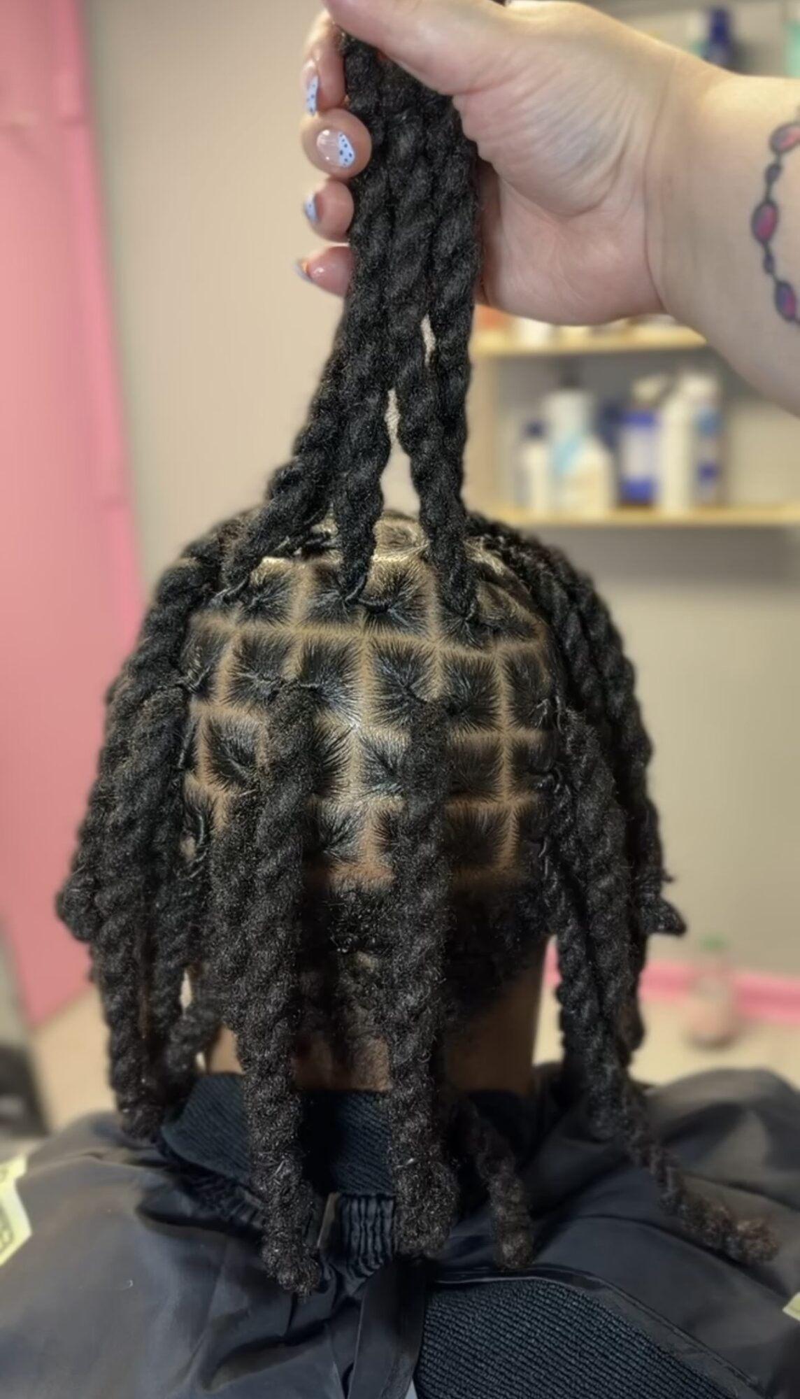 Person with styled dreadlocks, with a hand holding some of the hair, in a room with shelves and bottles in the background.