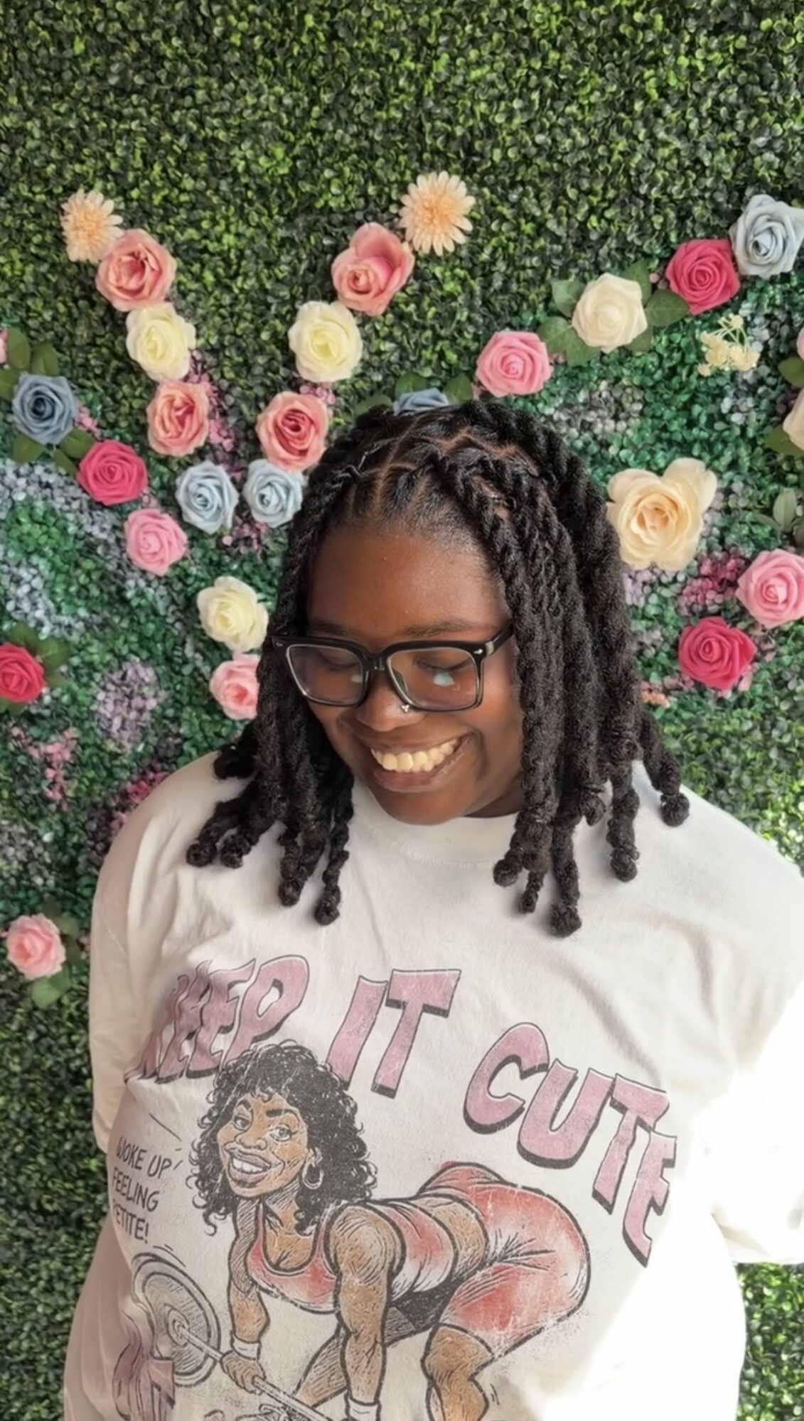 Smiling woman with glasses and dreadlocks in front of a colorful flower wall, wearing a white T-shirt with a cartoon character.