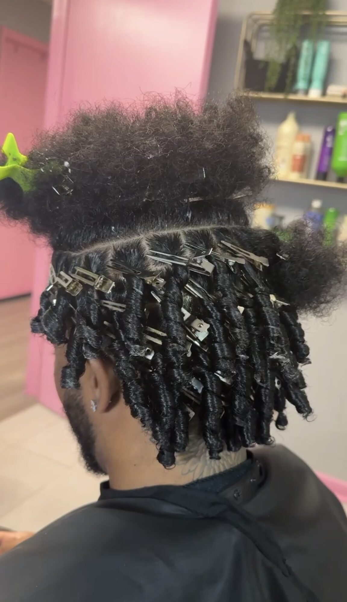 Person with styled dreadlocks and a fluffy afro hairstyle, sitting in a salon with shelves of hair products in the background.
