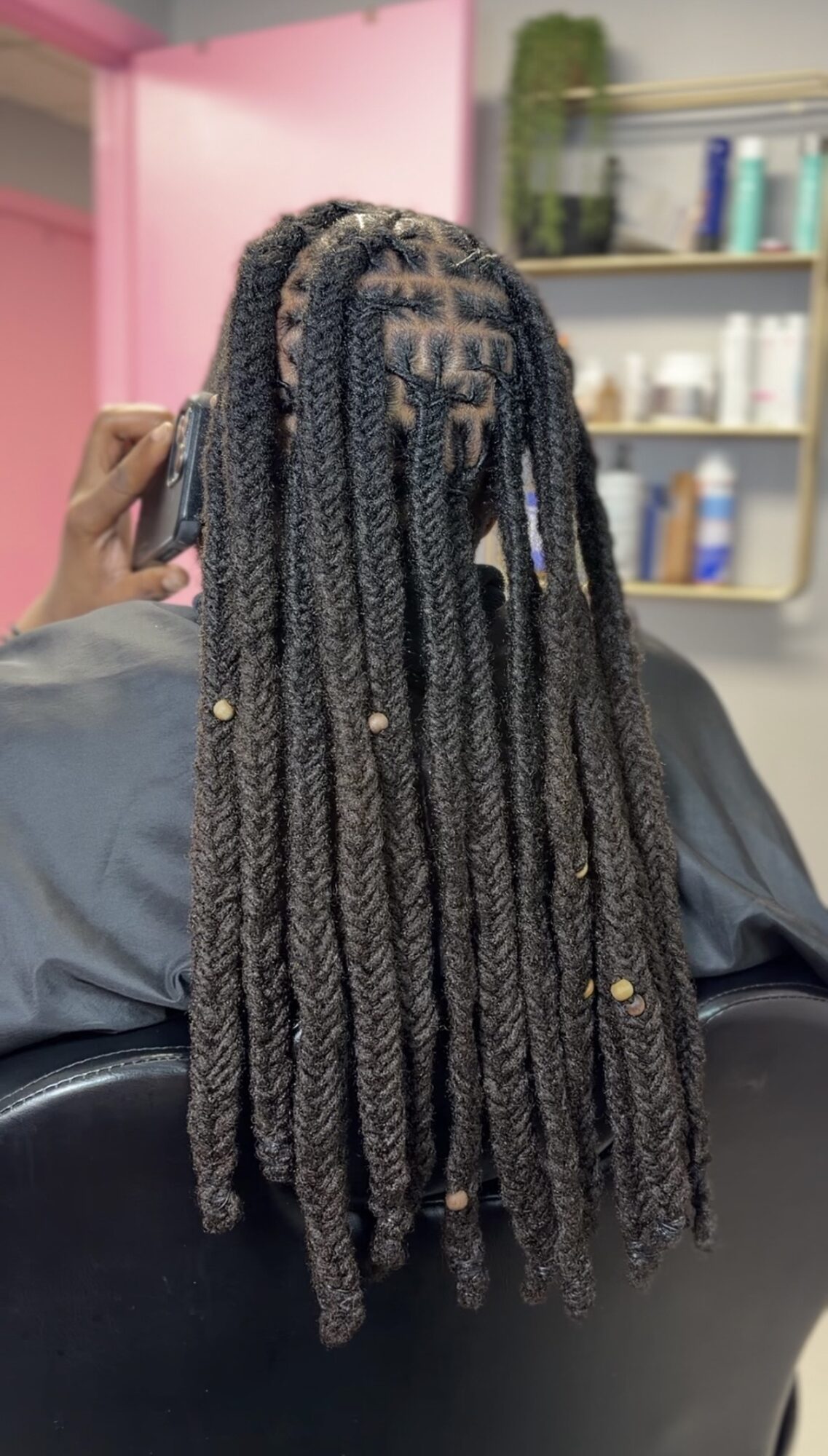 Back view of a person with long, gray dreadlocks in a salon chair, with shelves and pink wall in background.