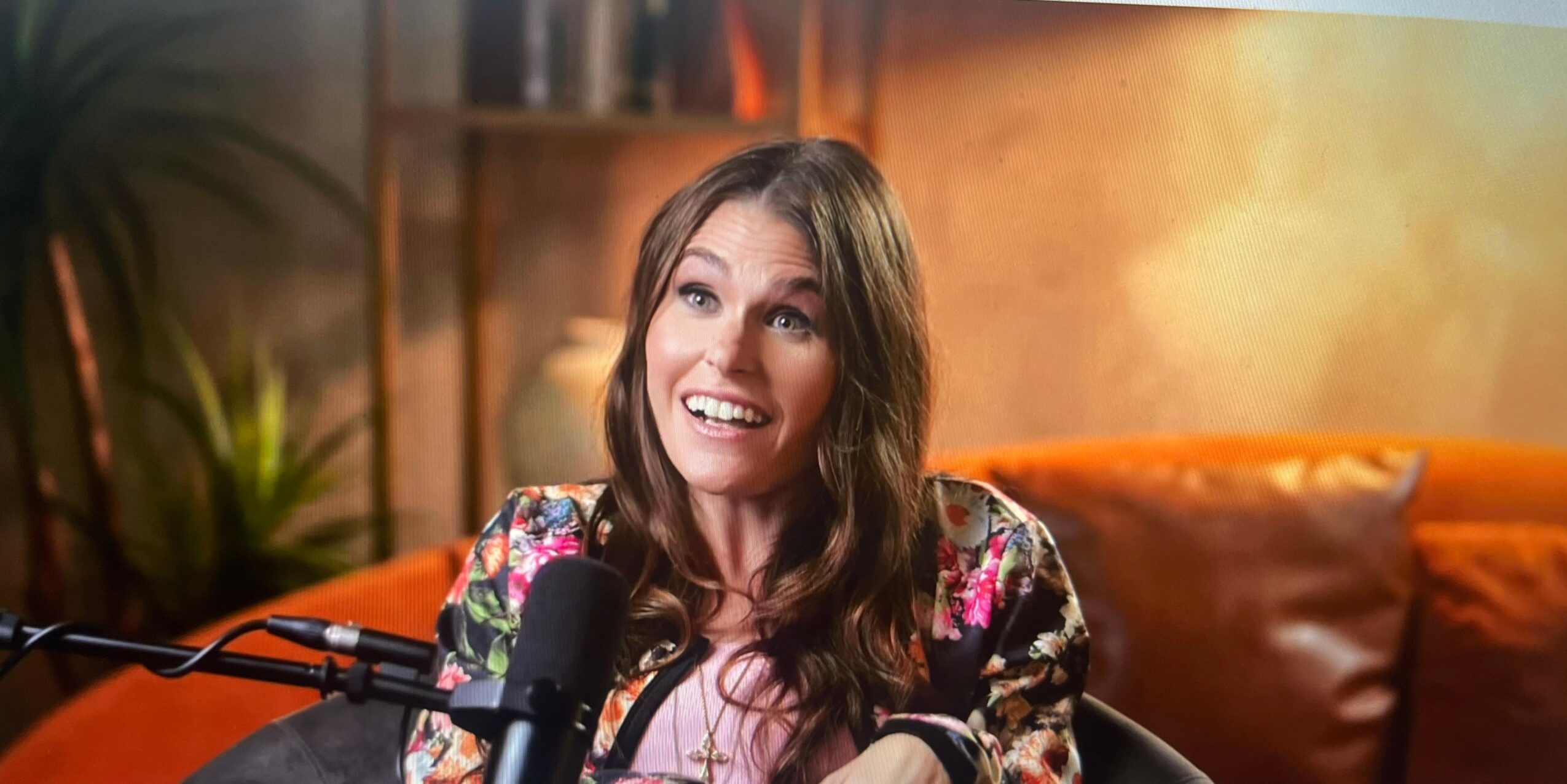 Young woman with long brown hair smiling, sitting in a cozy room with warm lighting and a microphone in front of her.