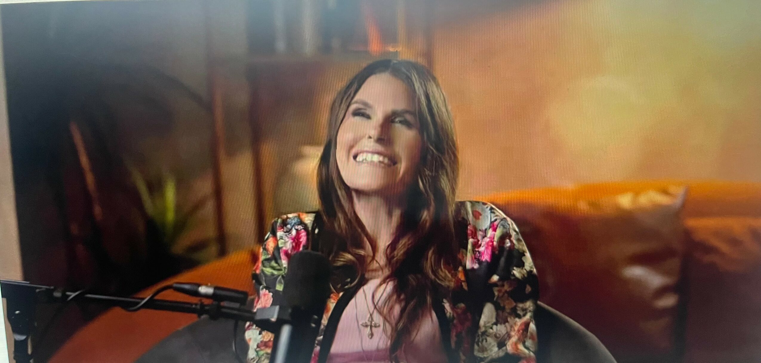 Woman smiling with long dark hair, wearing a floral top, in a warmly lit indoor setting.