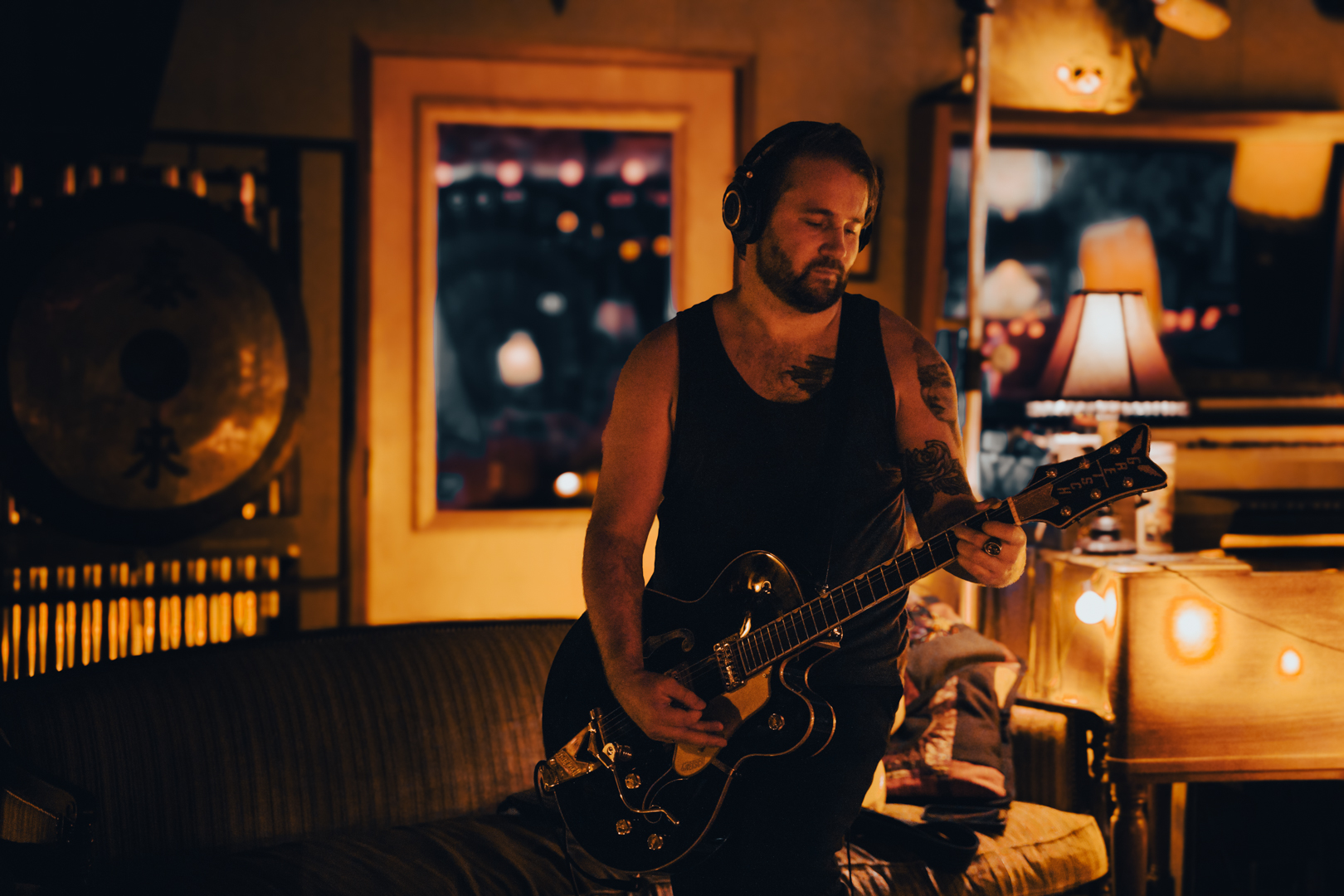 Man with tattoos playing an electric guitar in a warmly lit room at night, with a window and lamp in the background.