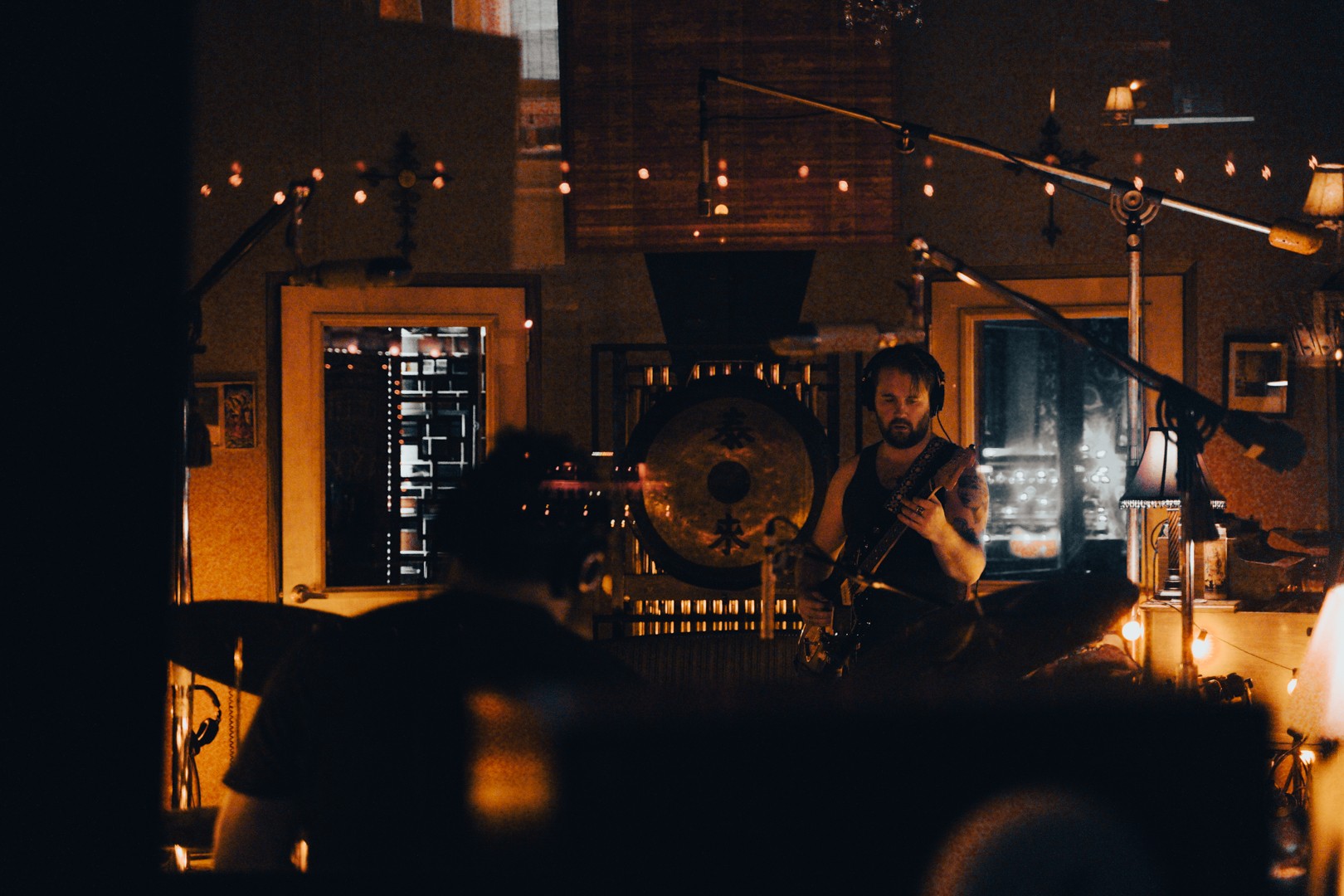 Musician playing a gong in a dimly lit room with warm lighting and string lights, with a person in the foreground.