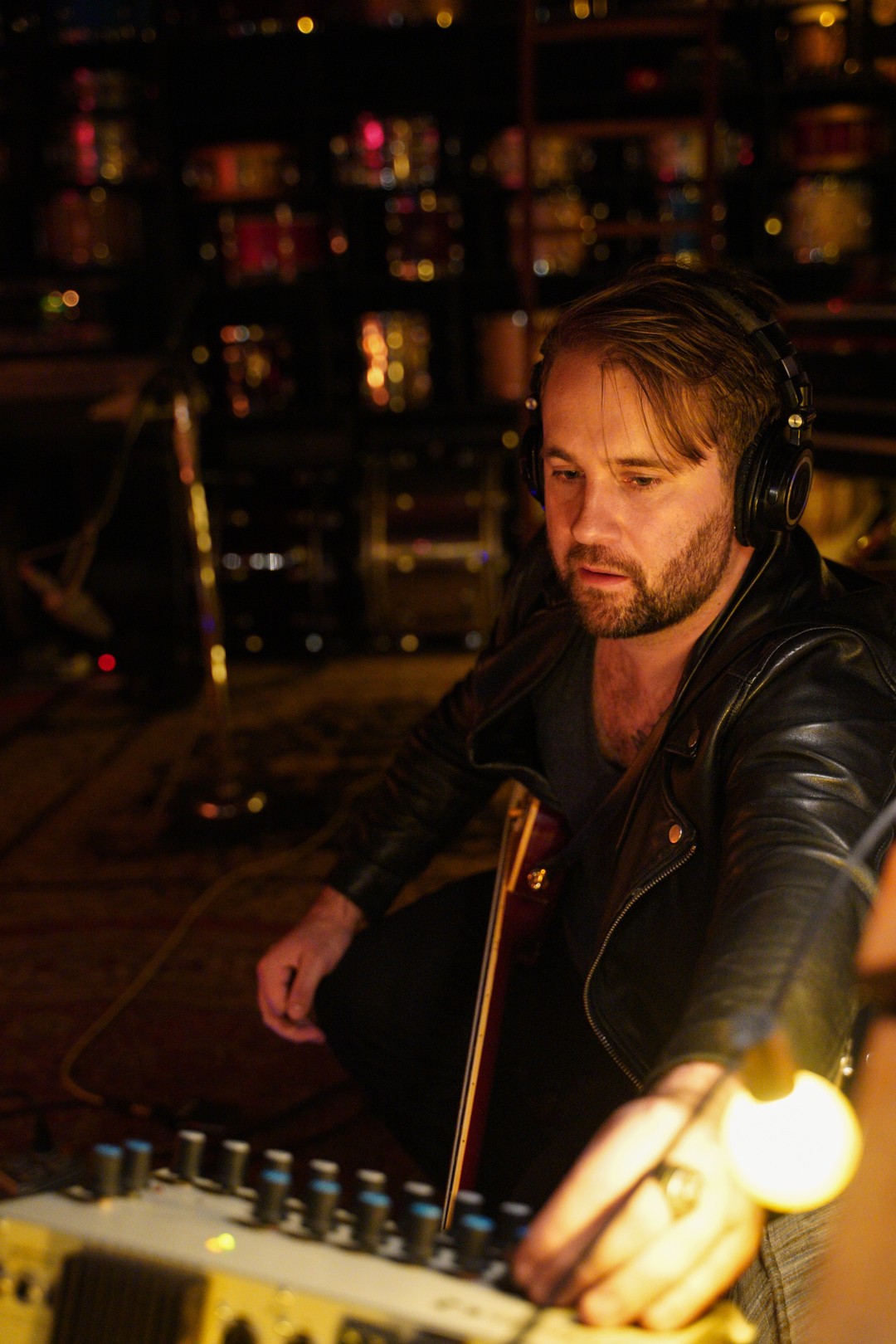 Man with headphones adjusting sound mixer in a dimly lit studio, shelves with equipment in background.