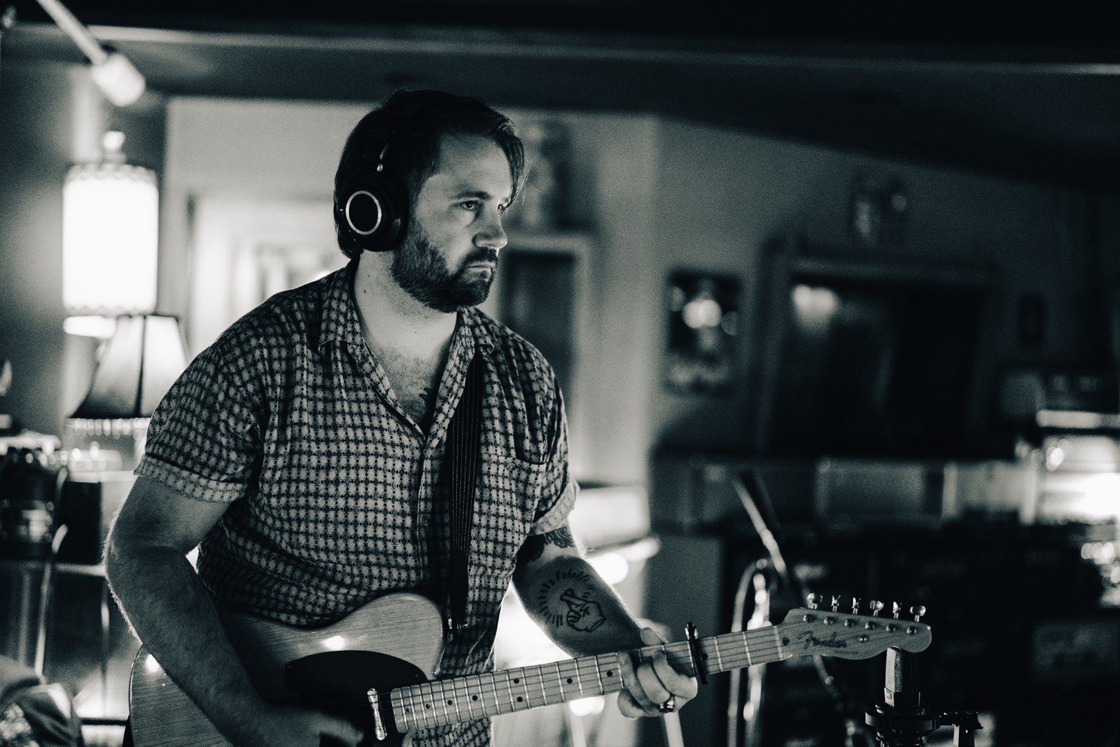 Man with headphones playing electric guitar in a dimly lit room, focused on music, with a tattoo on his arm.