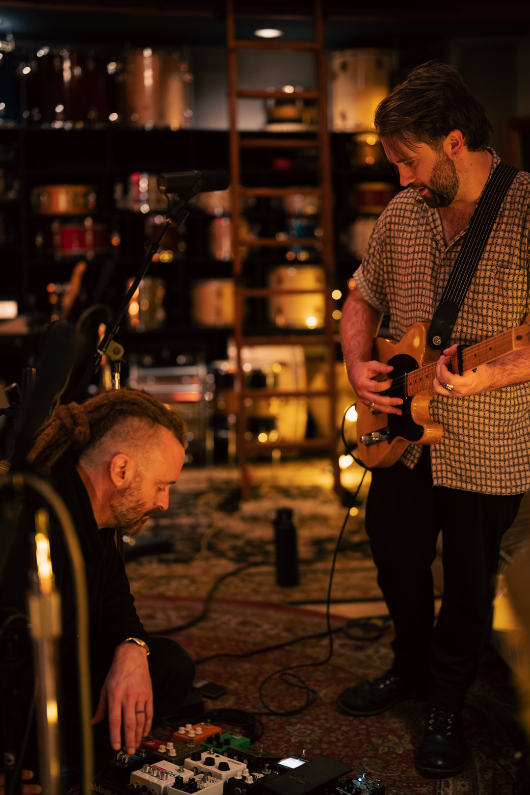 Two men playing guitar and using music equipment in a dimly lit room with shelves of jars or bottles in the background.