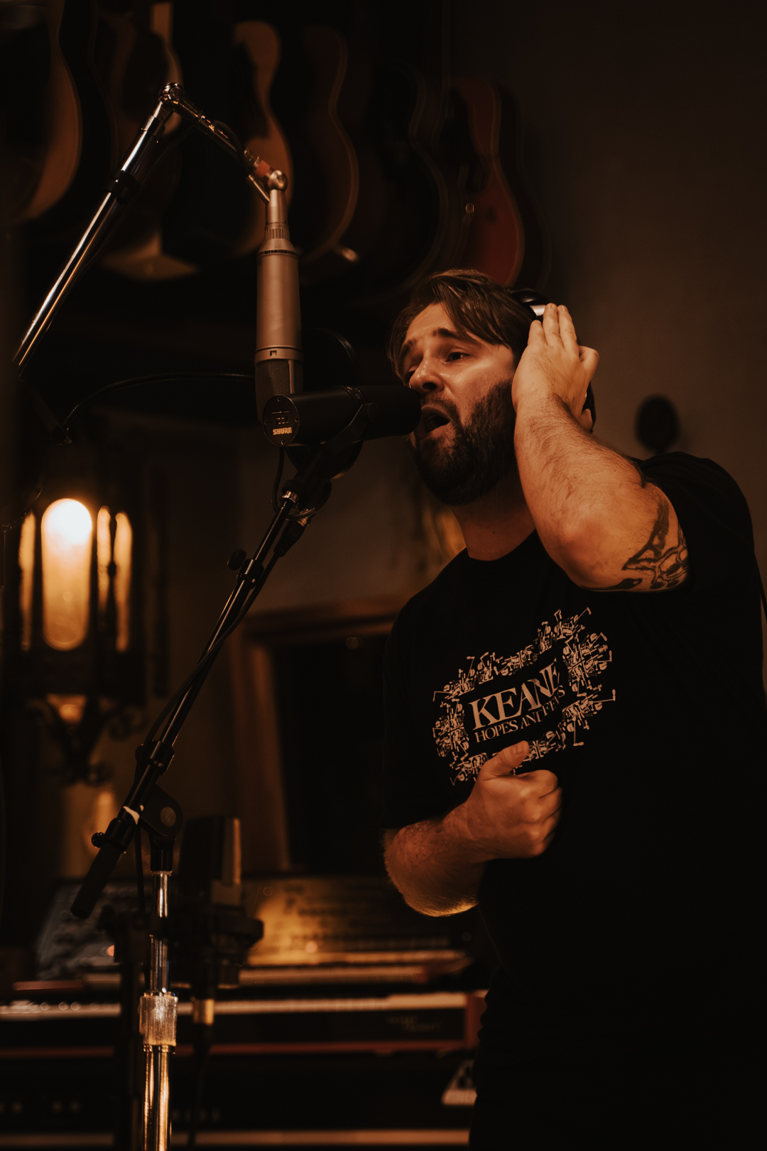 Man with beard singing into a microphone in a dimly lit room with guitars hanging on the wall behind him.
