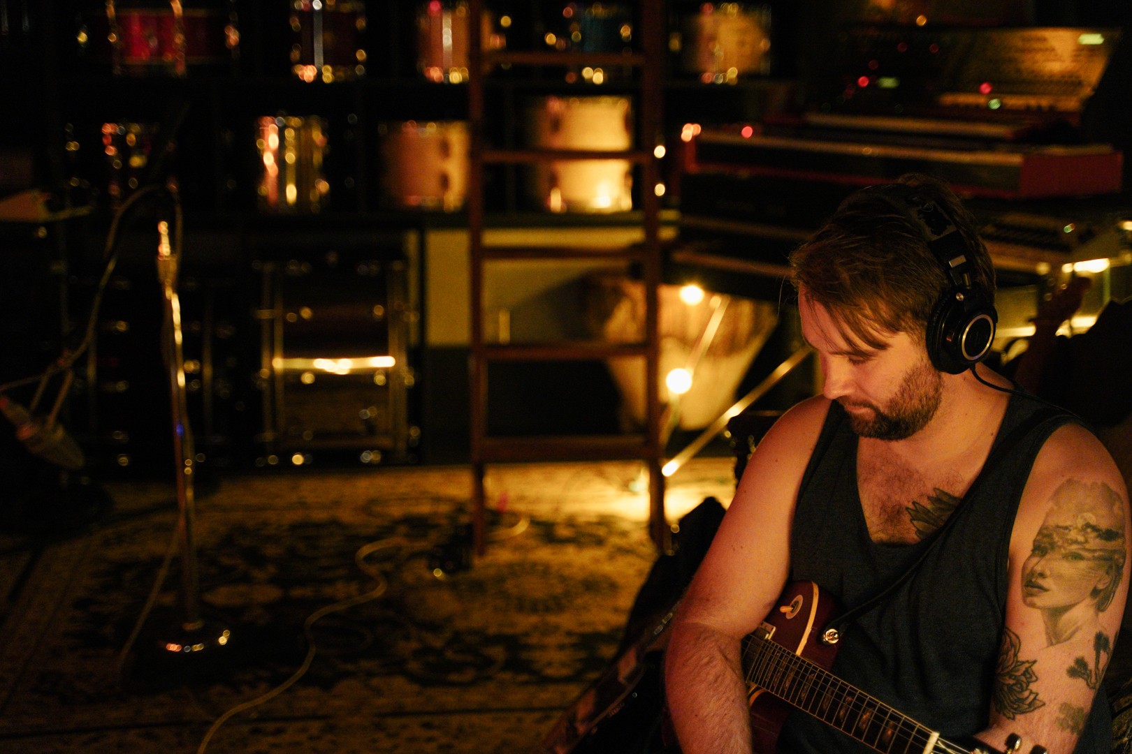 Man with headphones playing guitar in a dimly lit room with city lights outside window.