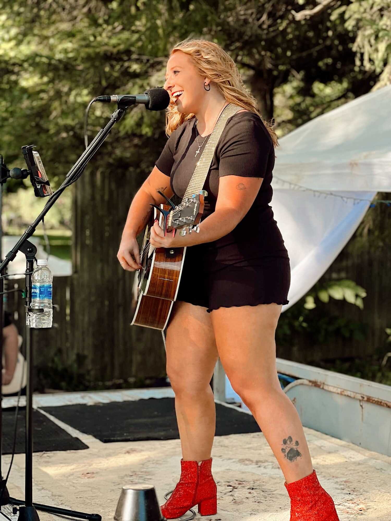 Woman with blonde hair singing into a microphone, playing guitar, outdoors with trees and a tent in background.