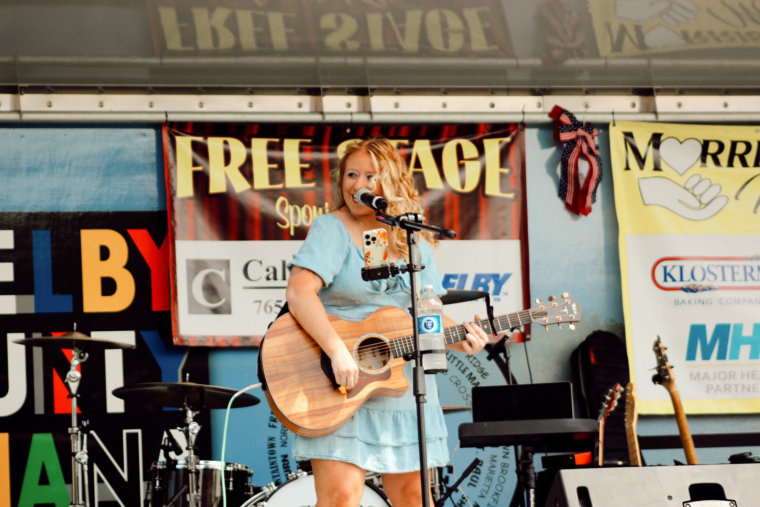 Young girl with blonde hair playing guitar and singing into a microphone on stage, wearing a light blue dress.