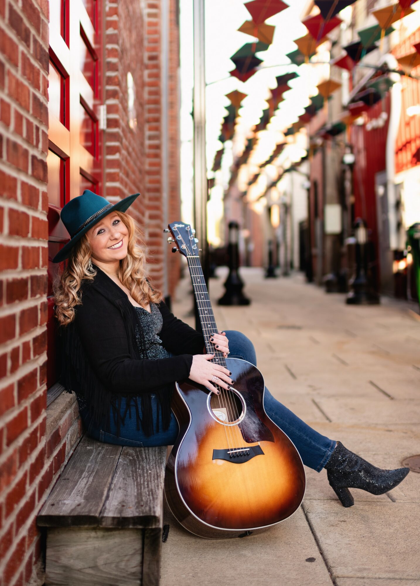 Woman with a guitar sitting on a bench in a city street, wearing a hat and black coat, smiling.
