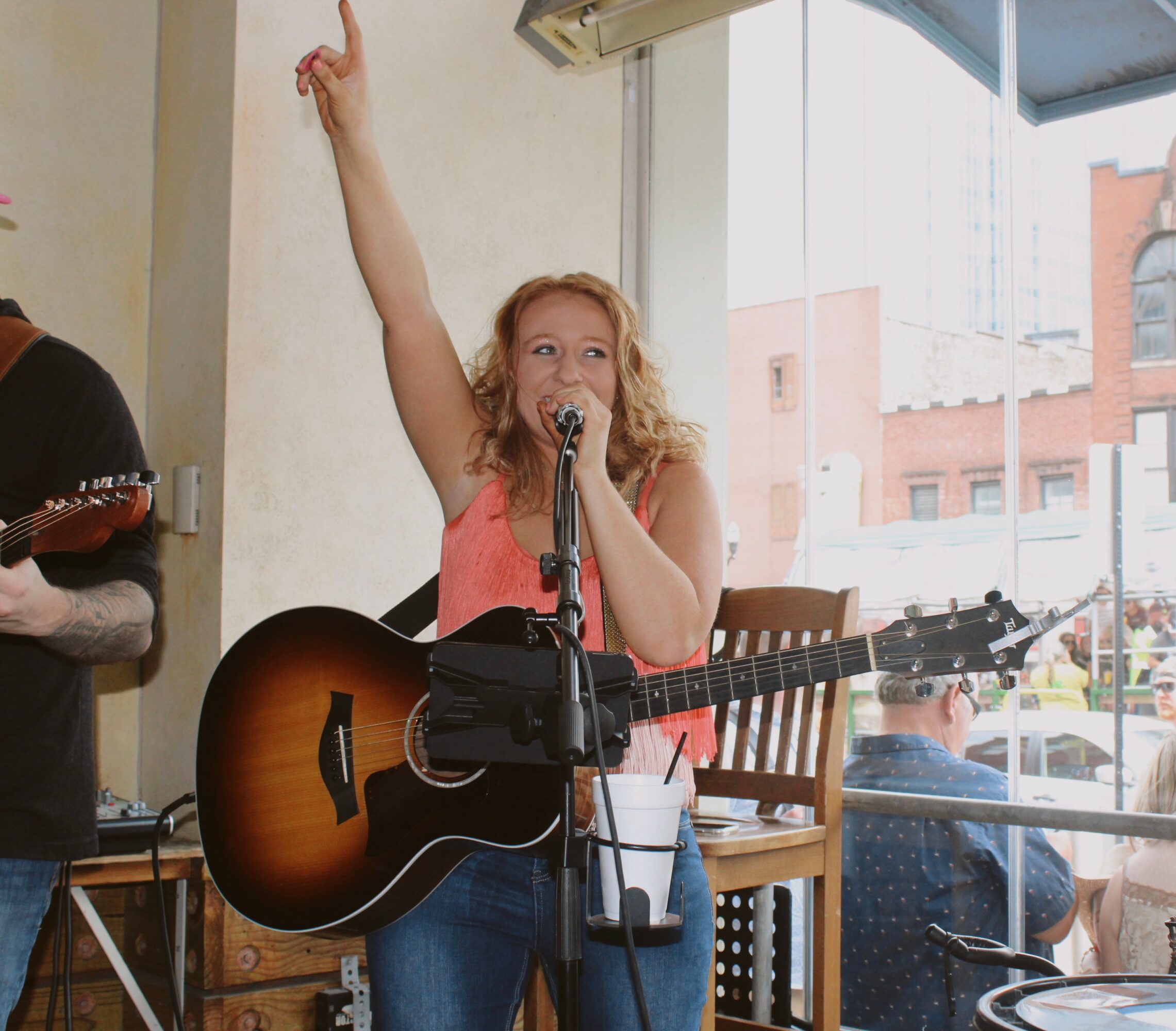Young woman singing into a microphone with her right arm raised, holding an acoustic guitar, in a cafe setting.