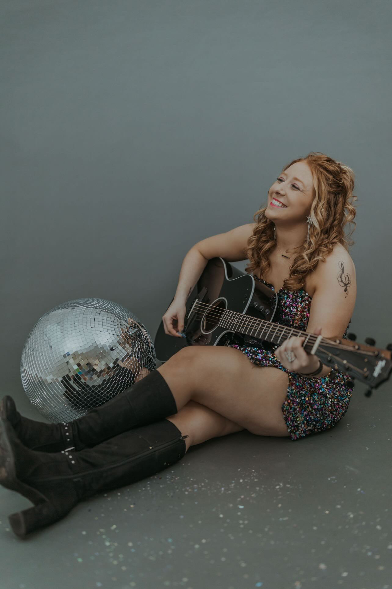 Young woman with curly hair sitting on the floor, playing guitar, smiling, with a disco ball nearby, against a plain background.