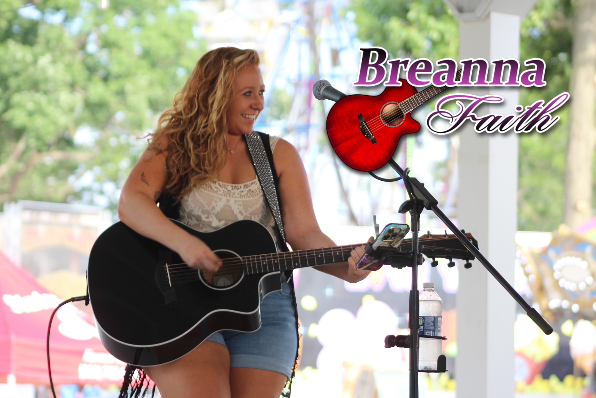 Woman with curly hair playing a black acoustic guitar on stage outdoors, with trees in background, and a logo reading 'Breanna Faith' with a guitar icon.