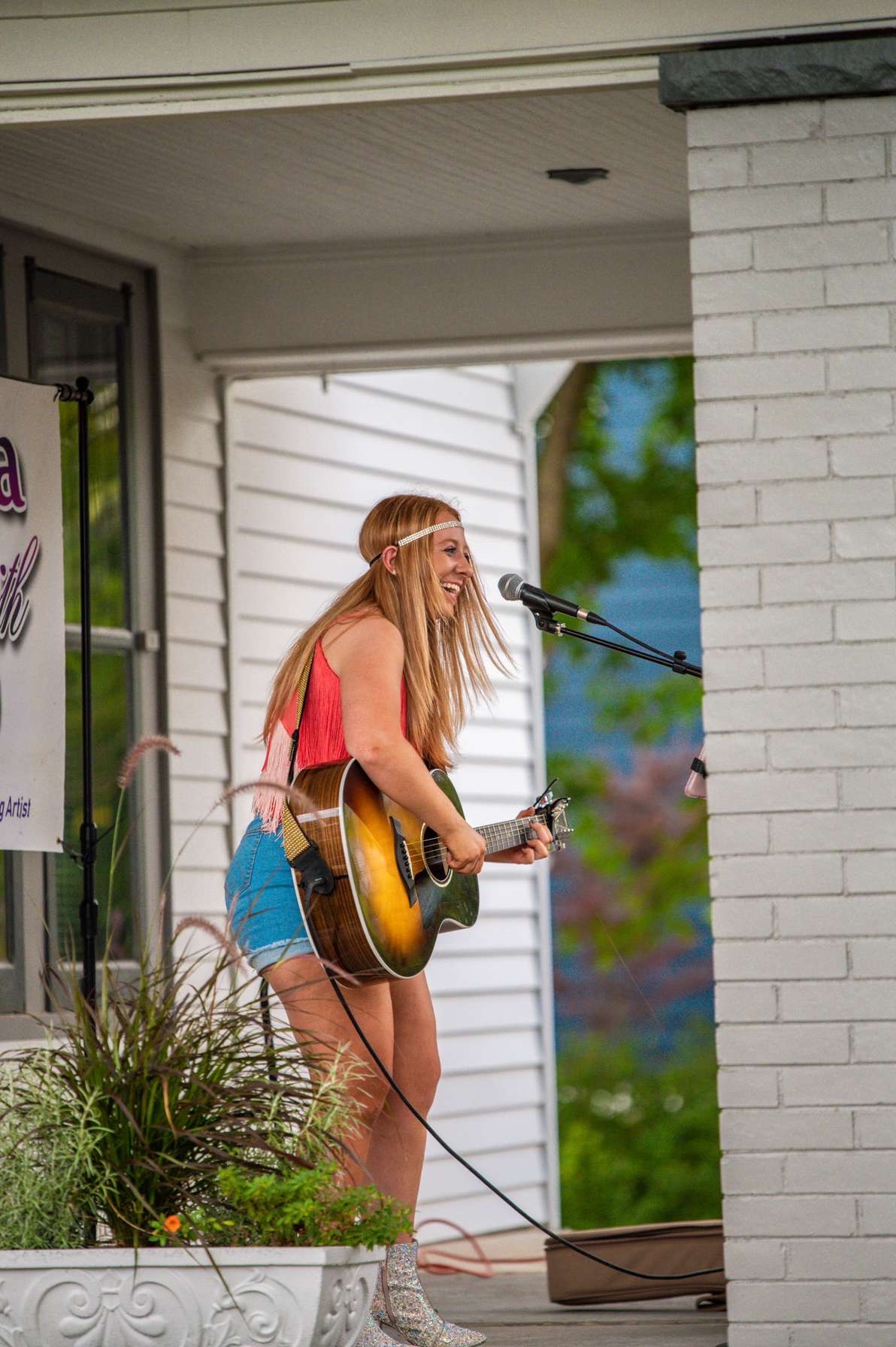 Young woman with long hair playing guitar and singing into a microphone outdoors, standing near a white brick wall.