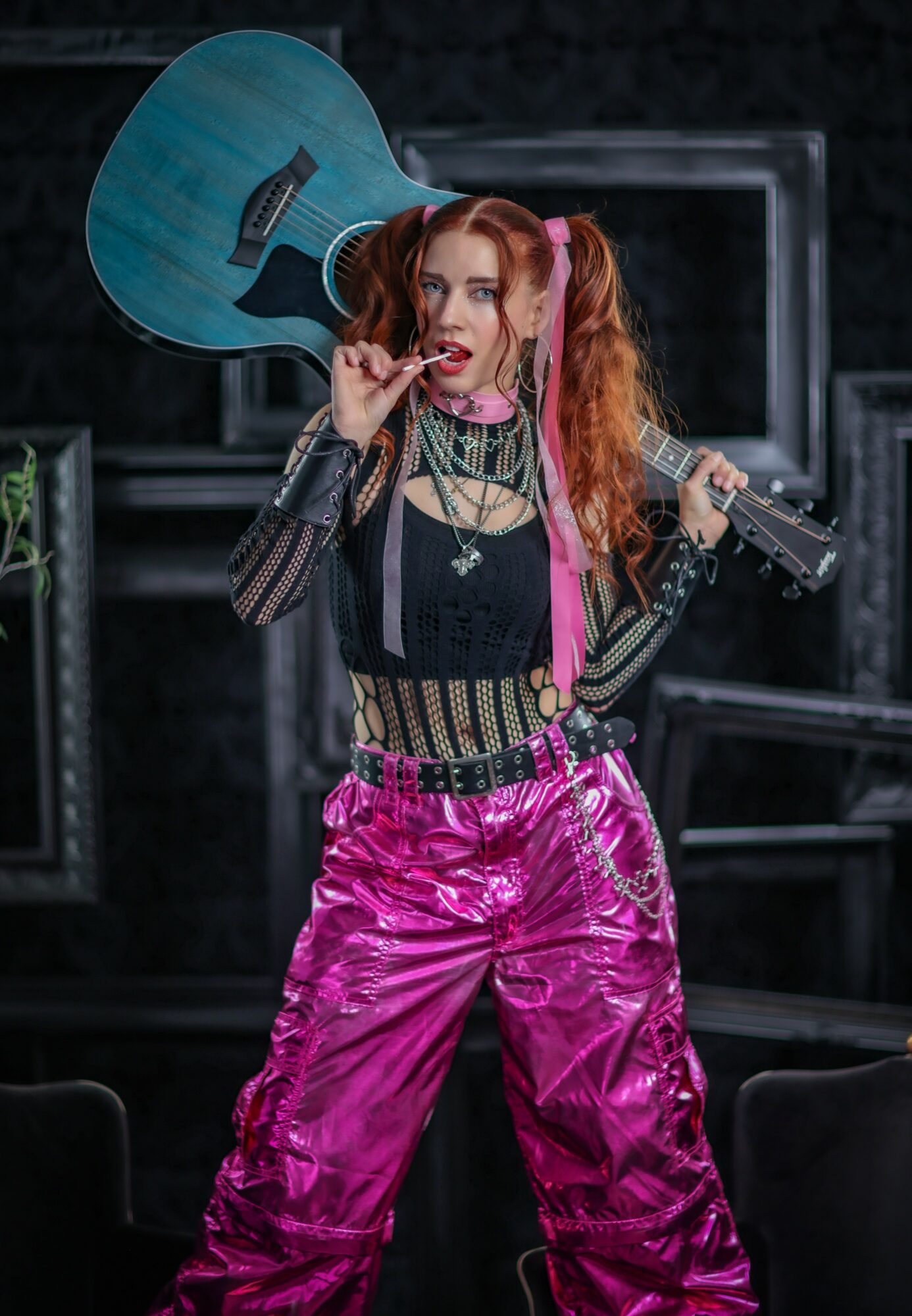 Young woman with colorful hair holding a guitar over her shoulder, wearing shiny pink pants and a black top, on stage.