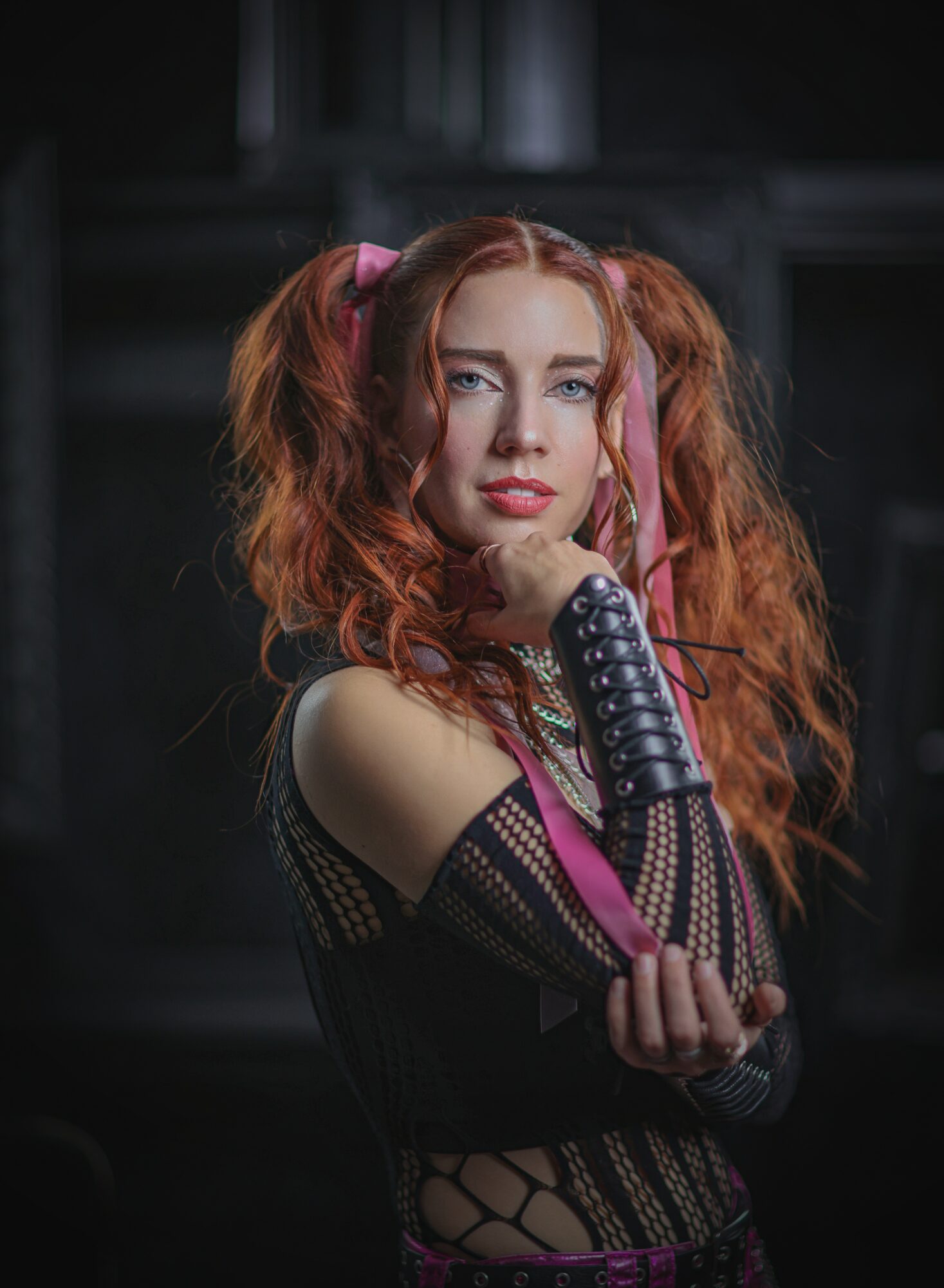 Young woman with curly red hair and pink hair accessories, wearing a black fishnet top, looking at the camera.