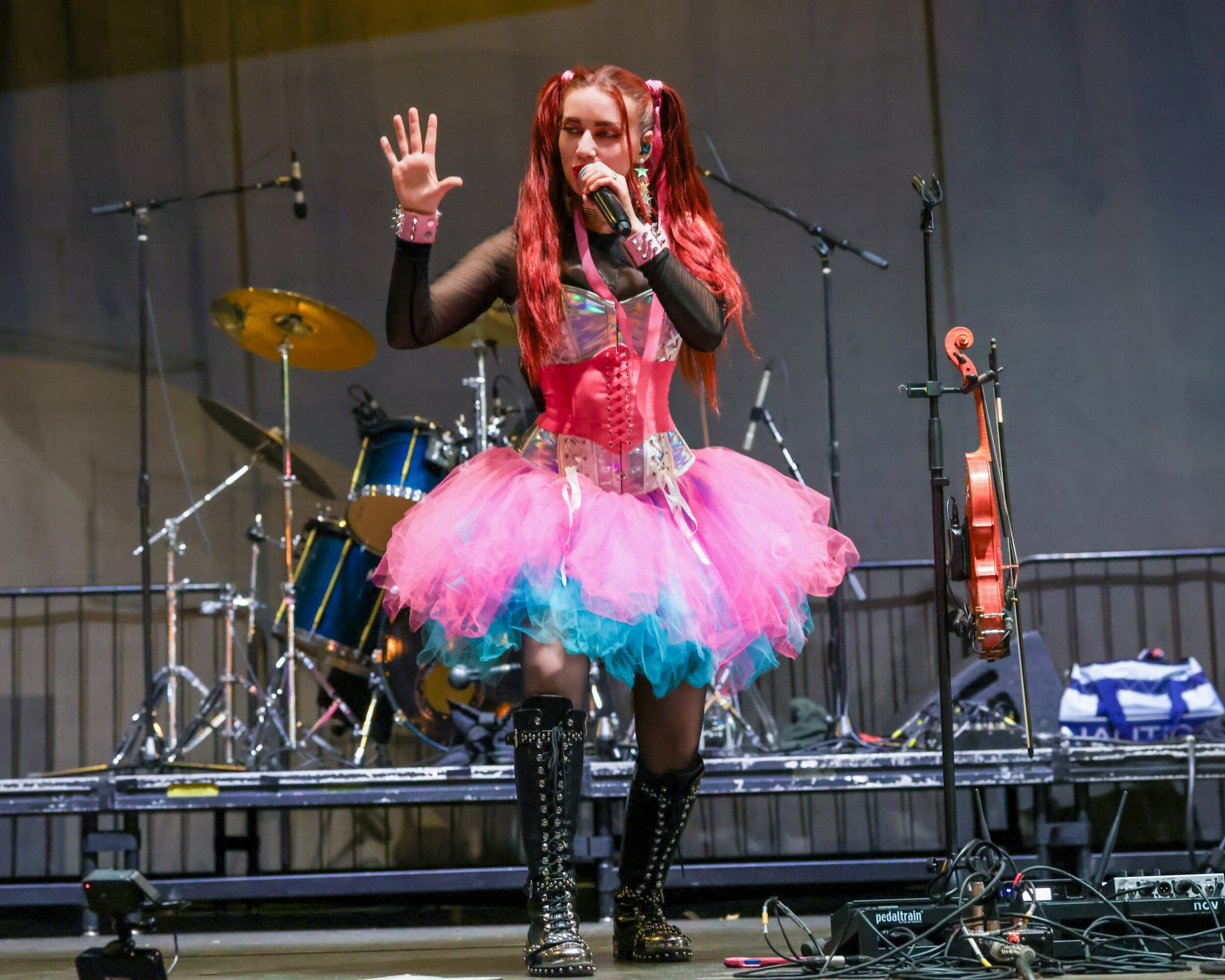 Performer with pink and blue tutu singing into a microphone on stage, with drums and a guitar in the background.