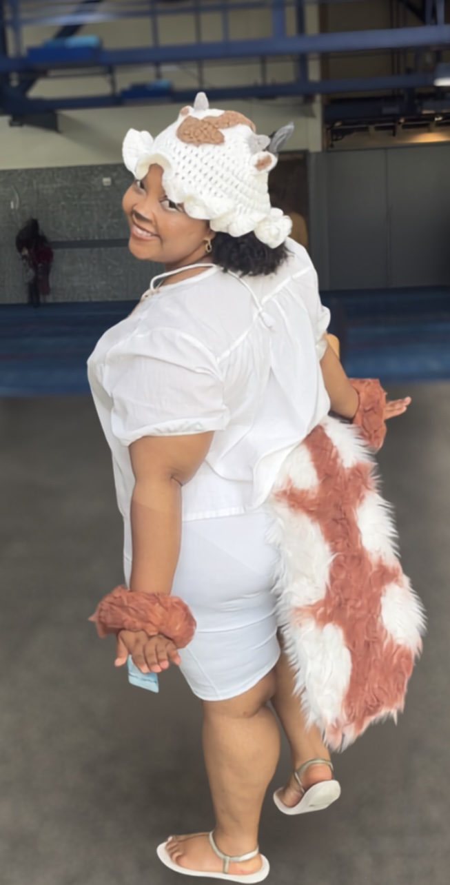 Person in a cow costume with a white outfit, cow ears, and a tail, standing indoors and smiling sideways.