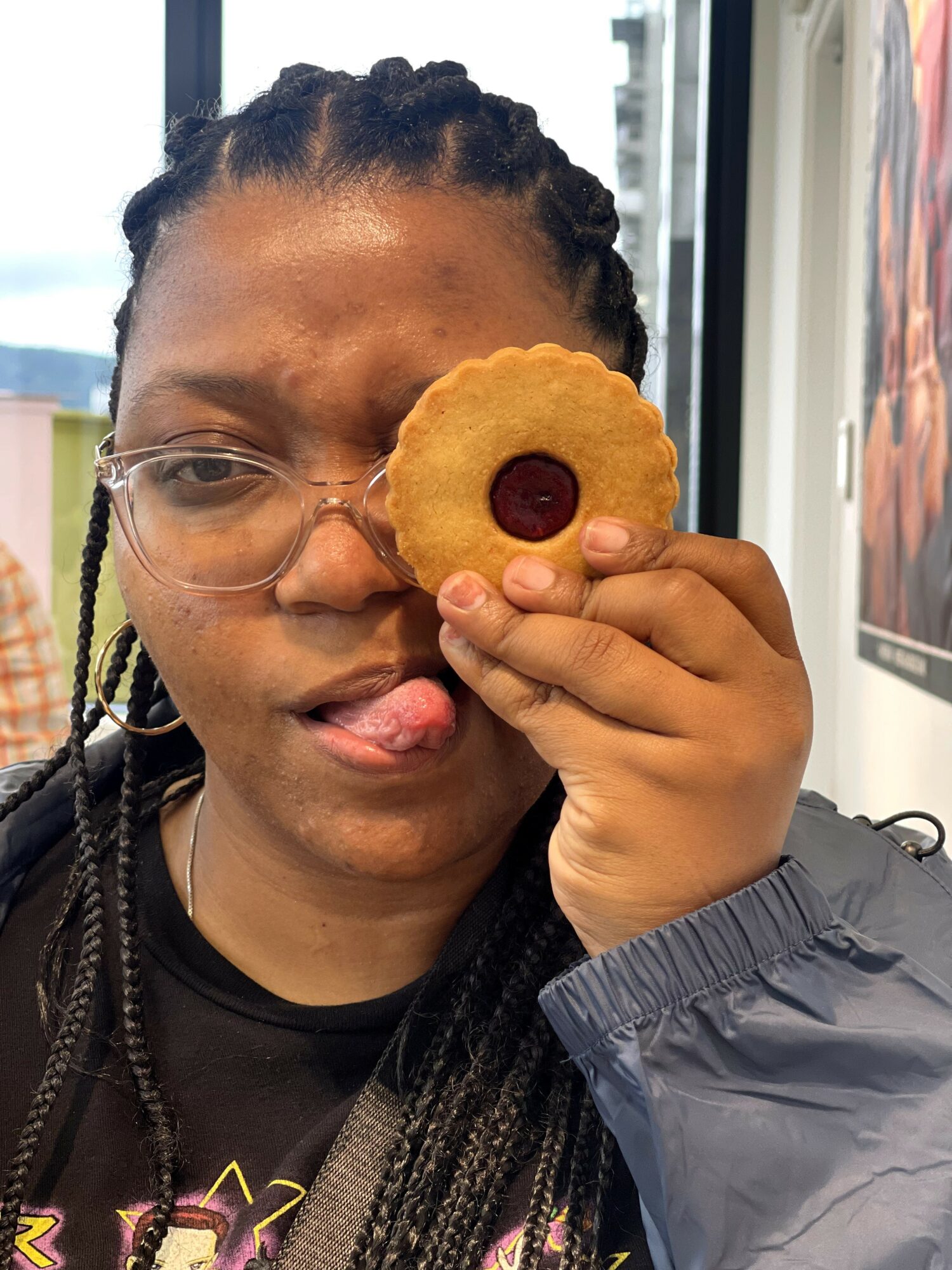Person holding a cookie with a red center, covering one eye, indoors with a window in the background.