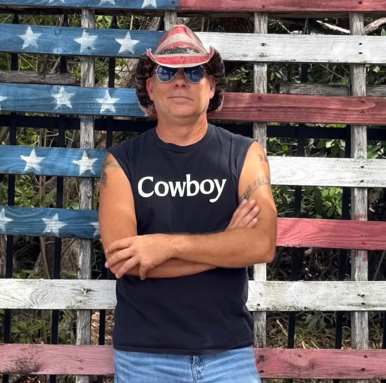 Person wearing sunglasses, cowboy hat, and sleeveless shirt with 'Cowboy' text, standing with arms crossed in front of a patriotic wooden background.