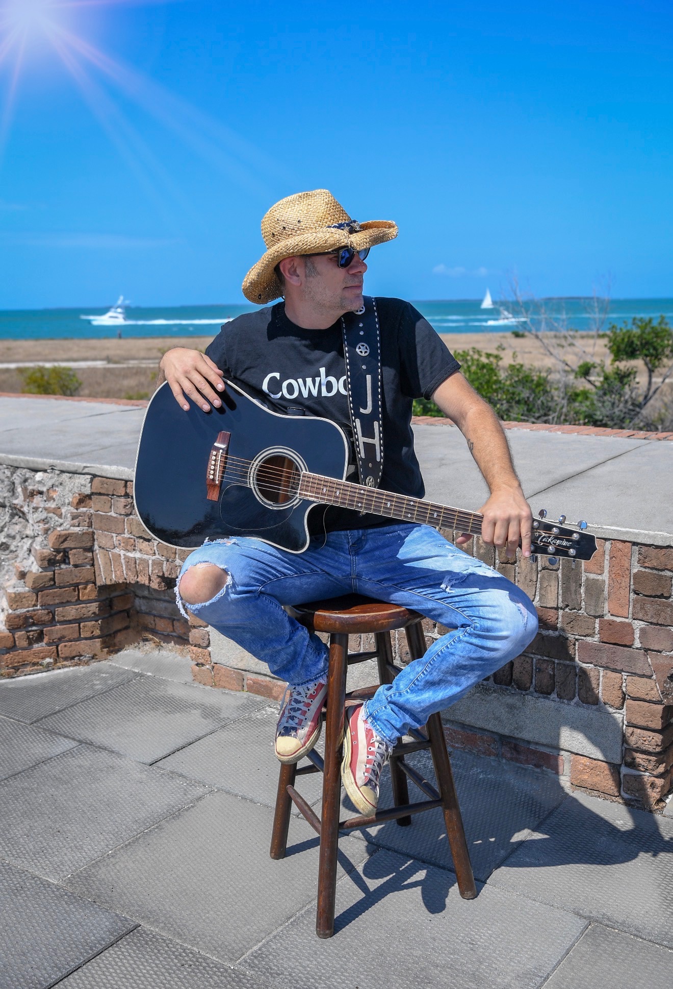 Person sitting on a stool outdoors holding an acoustic guitar, wearing a straw hat, sunglasses, and ripped jeans, with a beach and ocean in the background.