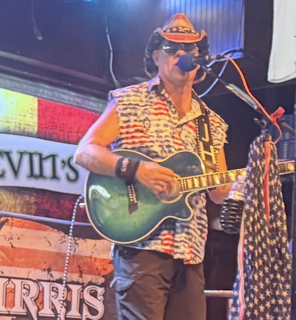 Person playing guitar on stage, wearing a hat, sunglasses, and a patterned sleeveless shirt, with a colorful background.