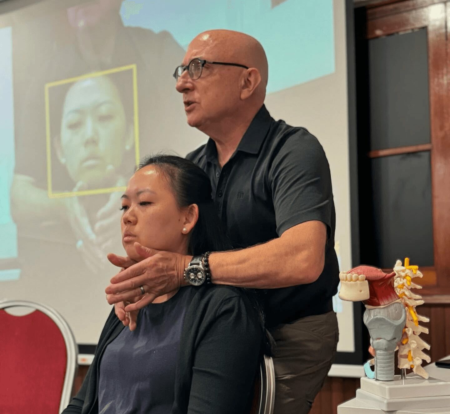 A man stands behind a seated woman, holding her shoulders, in a room with a presentation screen and a model of a spine.
