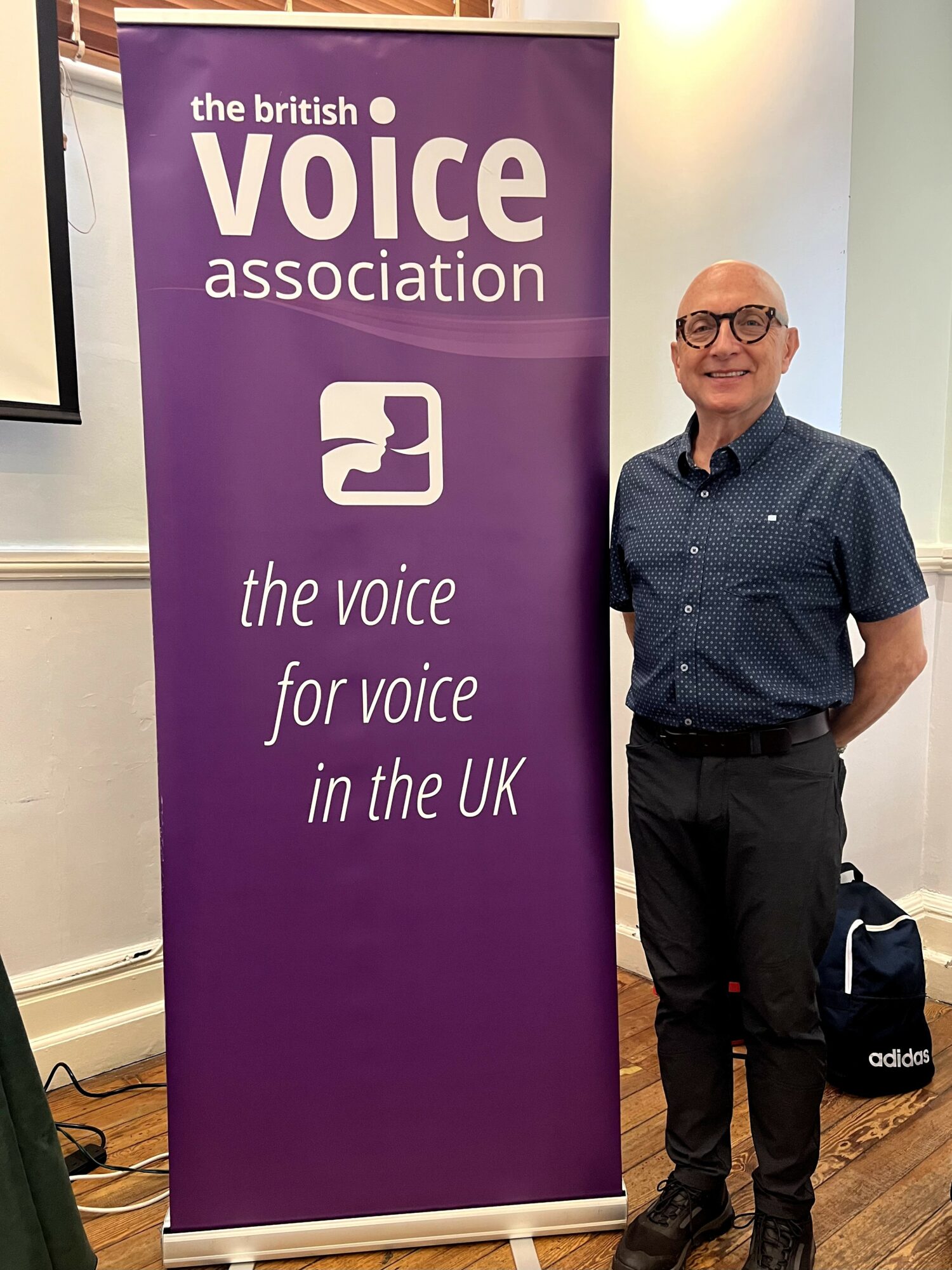 Man standing next to a purple banner for the British Voice Association, indoors with wooden floor.