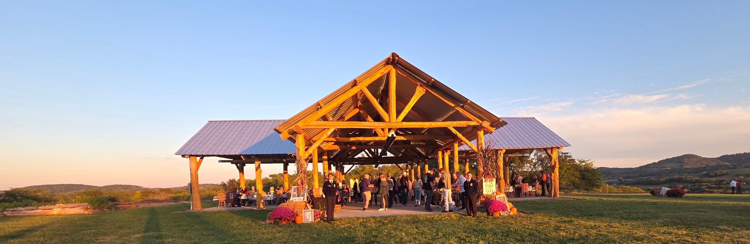 Open wooden pavilion with people gathered inside, set in a grassy landscape with hills in the background, during sunset.