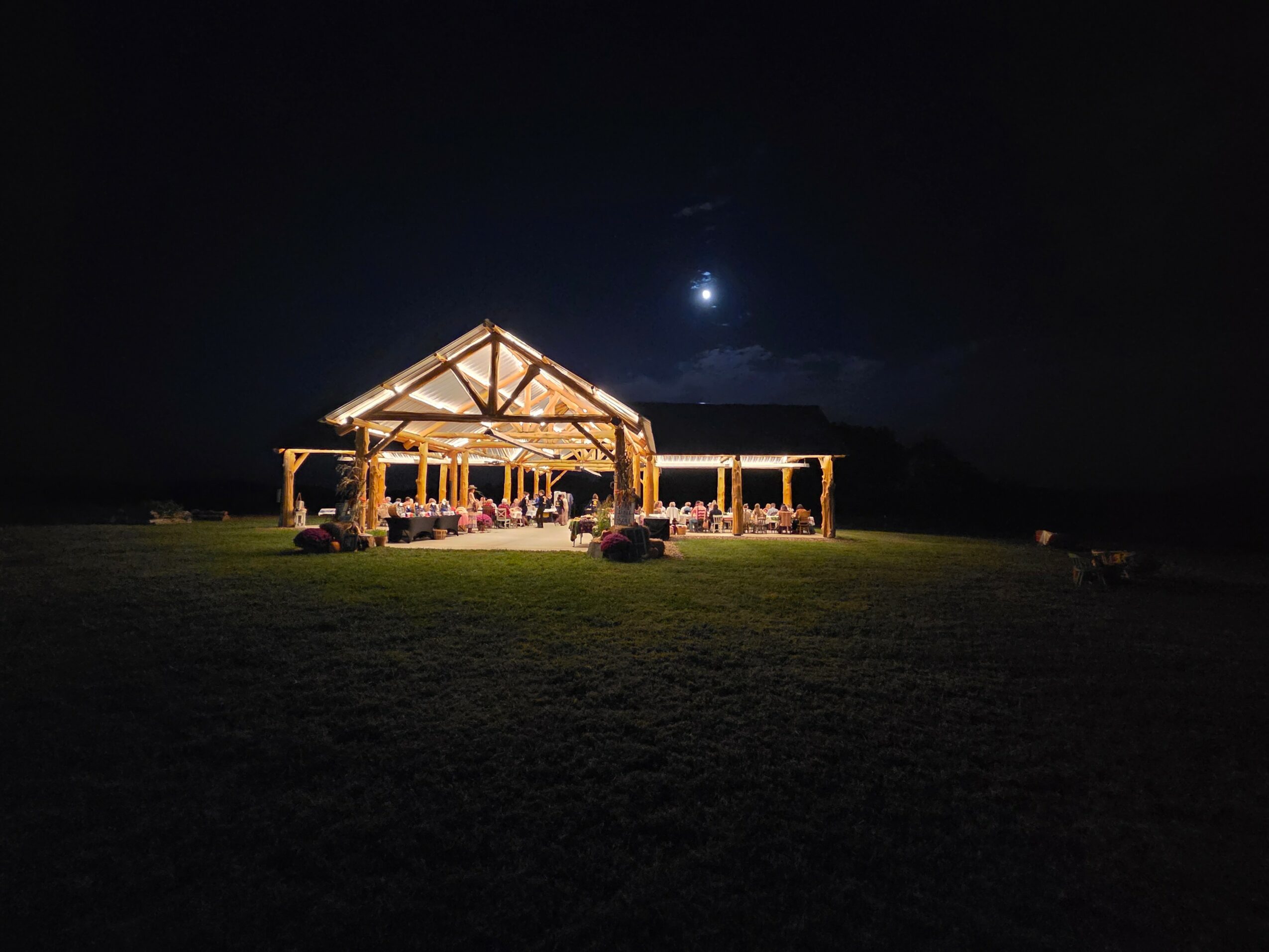 Open wooden pavilion illuminated at night with tables and chairs, moon visible in dark sky, grassy area in foreground.