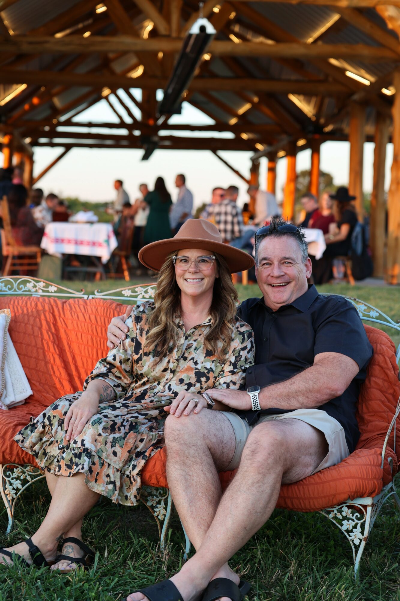 Two people sitting on a sofa outdoors, smiling, with a wooden structure and people in the background.