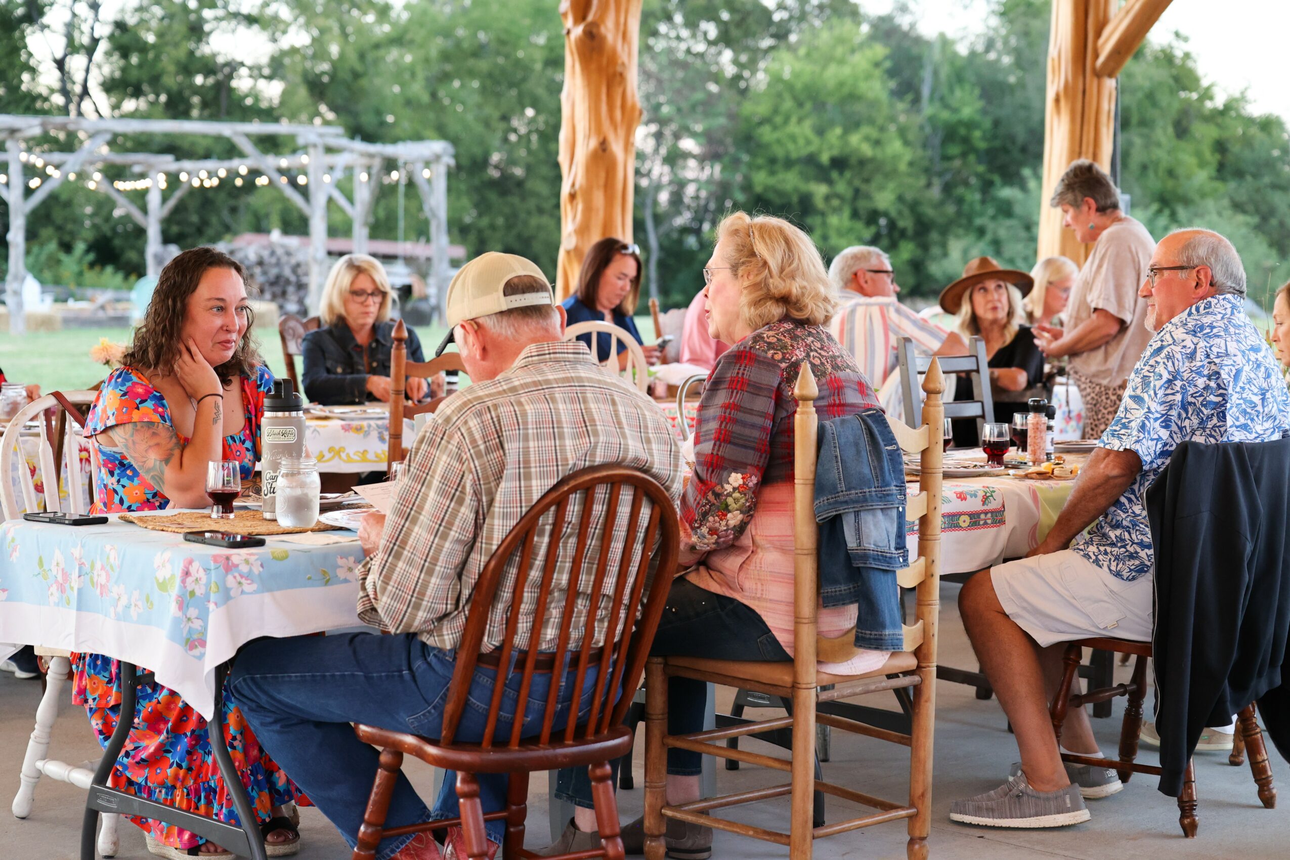 Group of people sitting at a long outdoor table with drinks, under a wooden structure, surrounded by trees.
