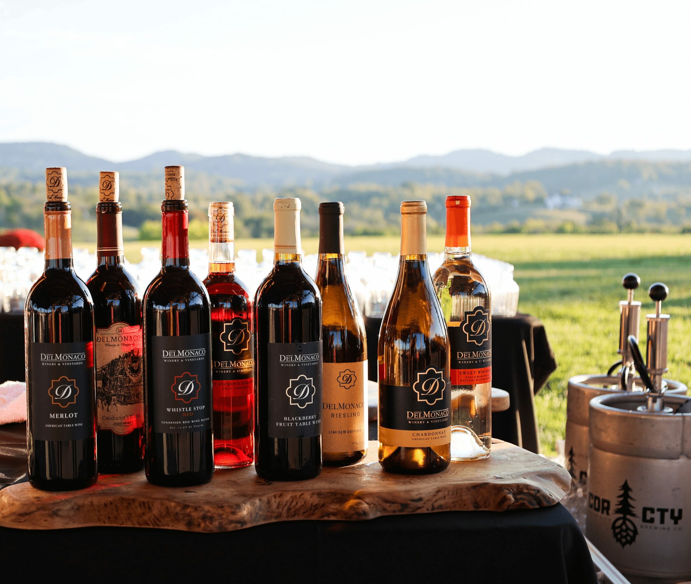Seven bottles of wine and a bottle opener on a wooden table outdoors with a landscape background.