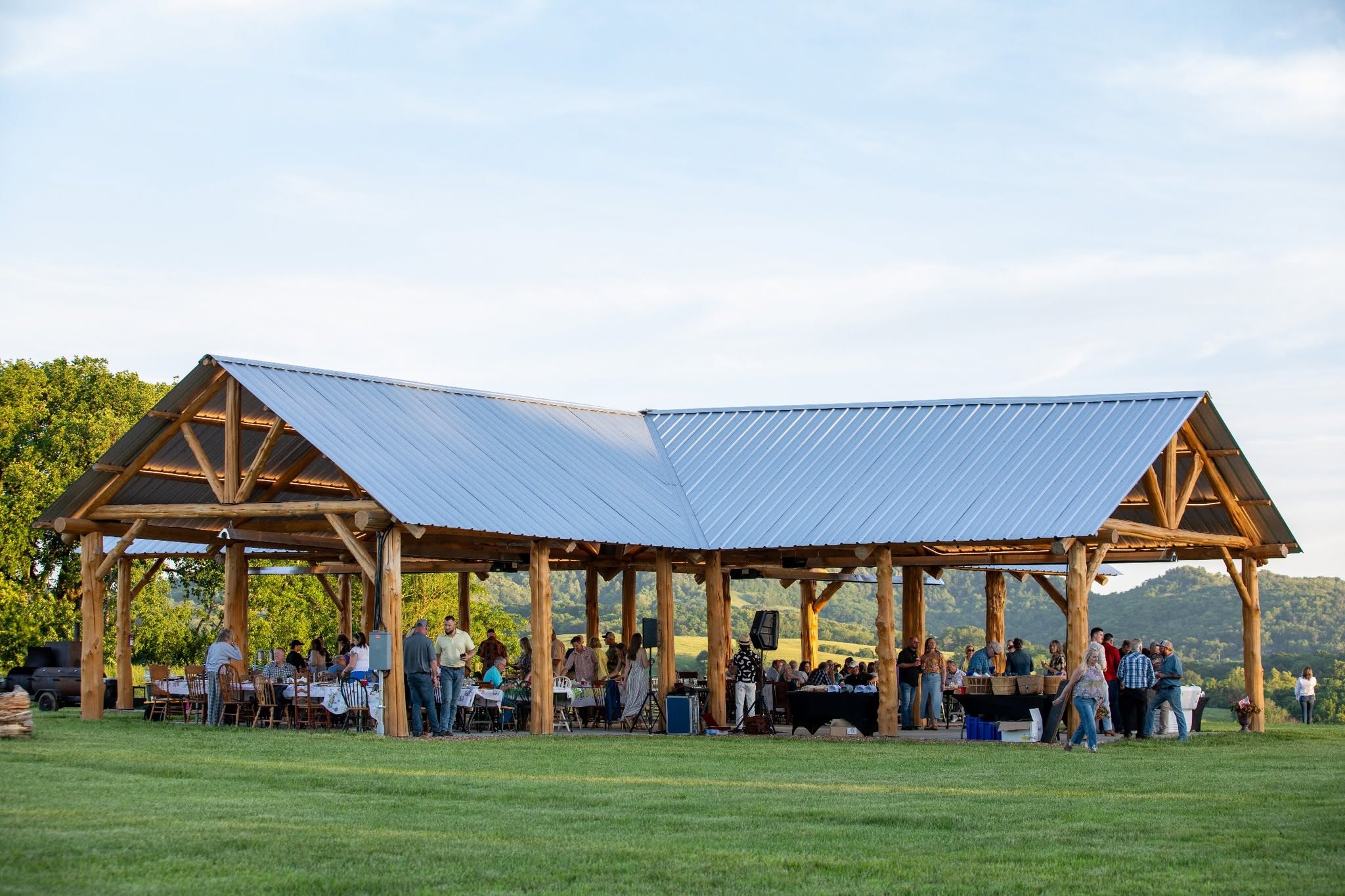 Open wooden pavilion with a blue metal roof, people dining outdoors on a grassy area, trees in the background.