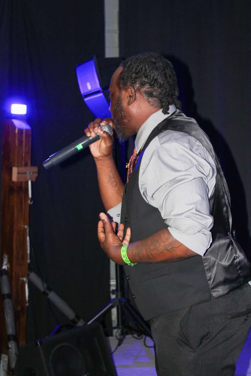 Person singing into a microphone on stage, wearing a white shirt and black vest, with dark background and stage lights.