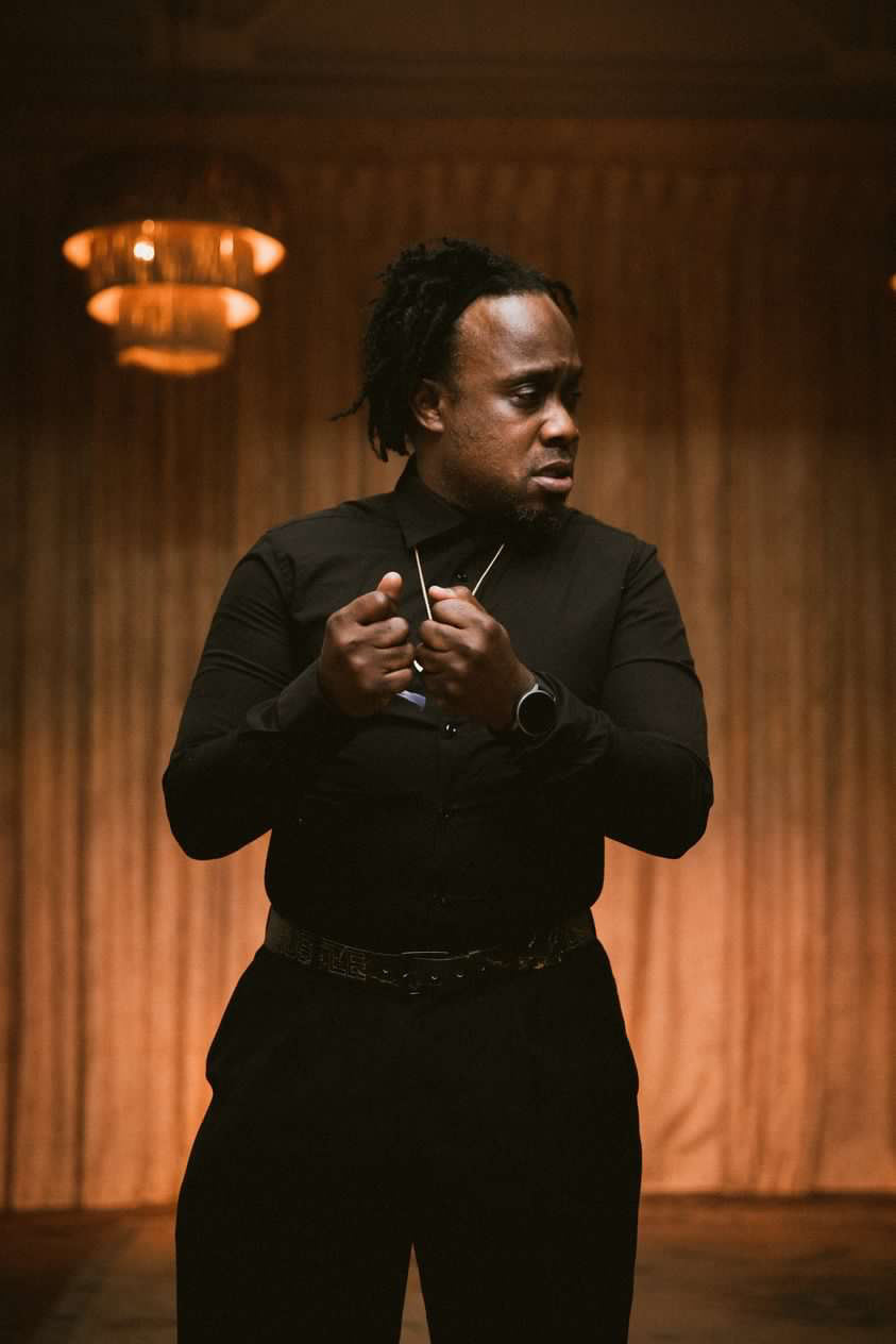 Person with dreadlocks wearing black clothing, holding glasses, standing in a room with wooden walls and warm lighting.