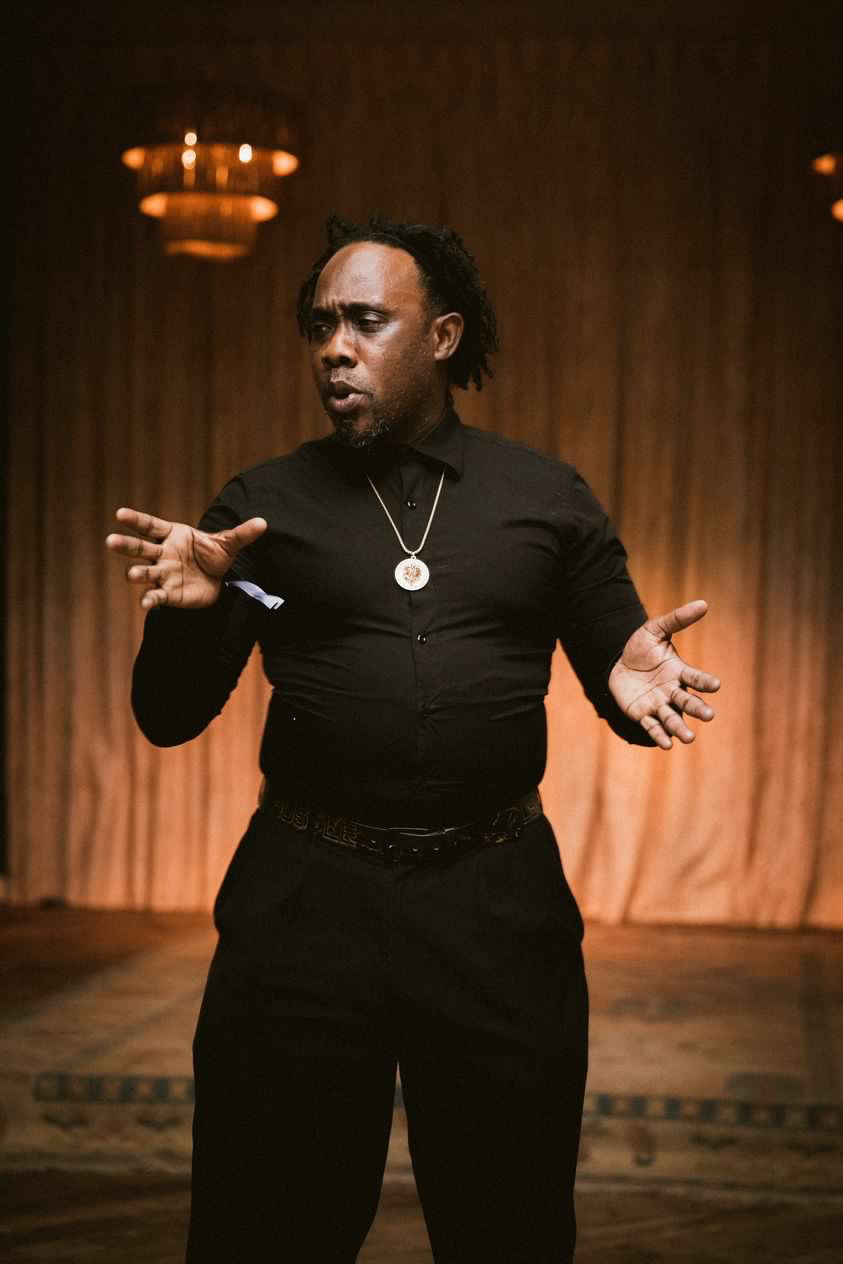 Person standing on stage with a curtain background, gesturing with hands, wearing black clothing and a necklace.