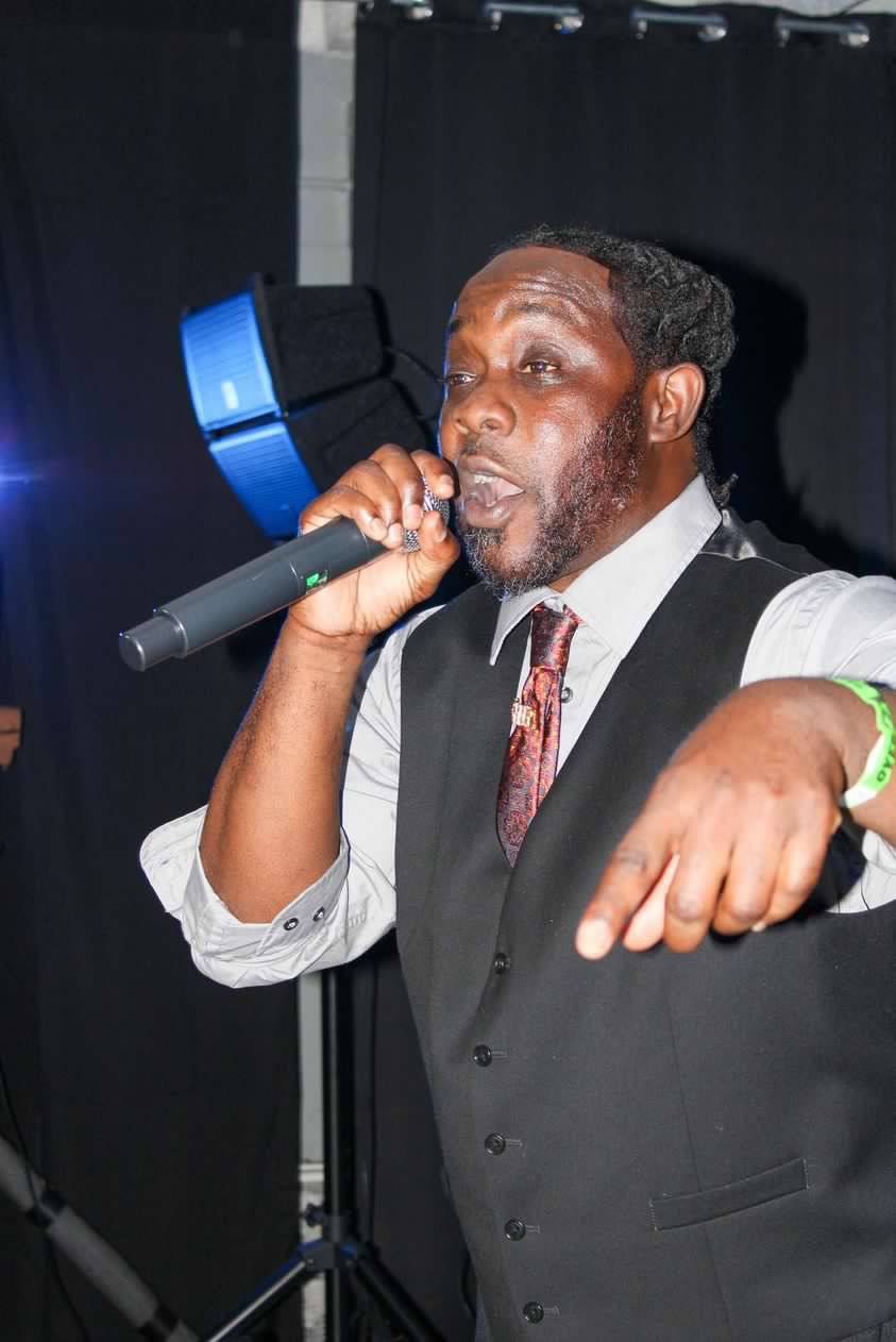 Man speaking into a microphone at an event, wearing a vest and tie, with a dark background.