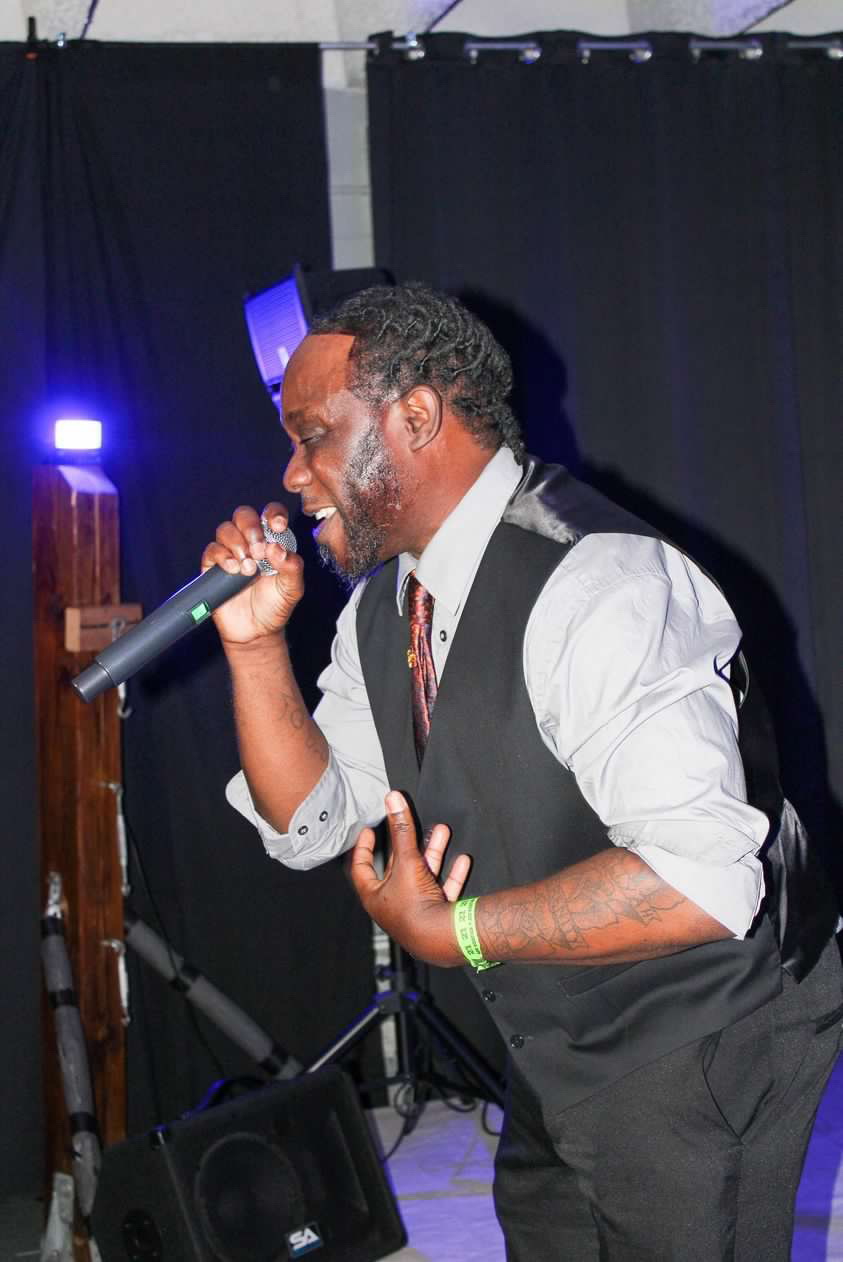 Man singing into microphone on stage, wearing a vest and white shirt, with black curtains in background.